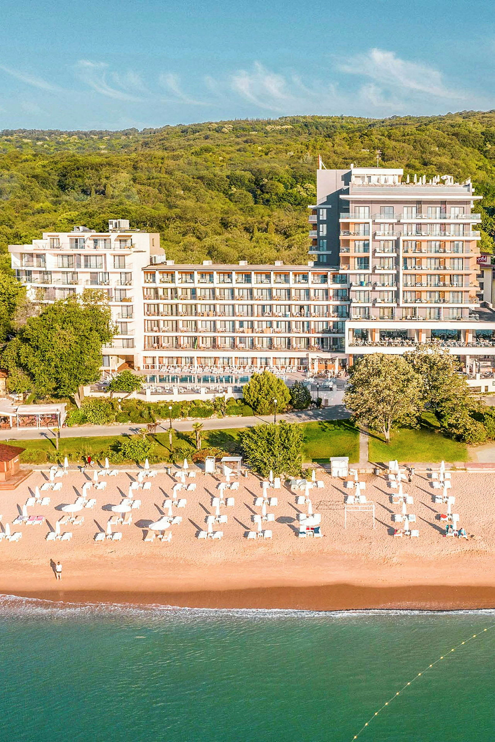 Udsigt over en strand med liggestole og parasoller ved et hotel, omgivet af grønne områder. (AI-genereret tekst)