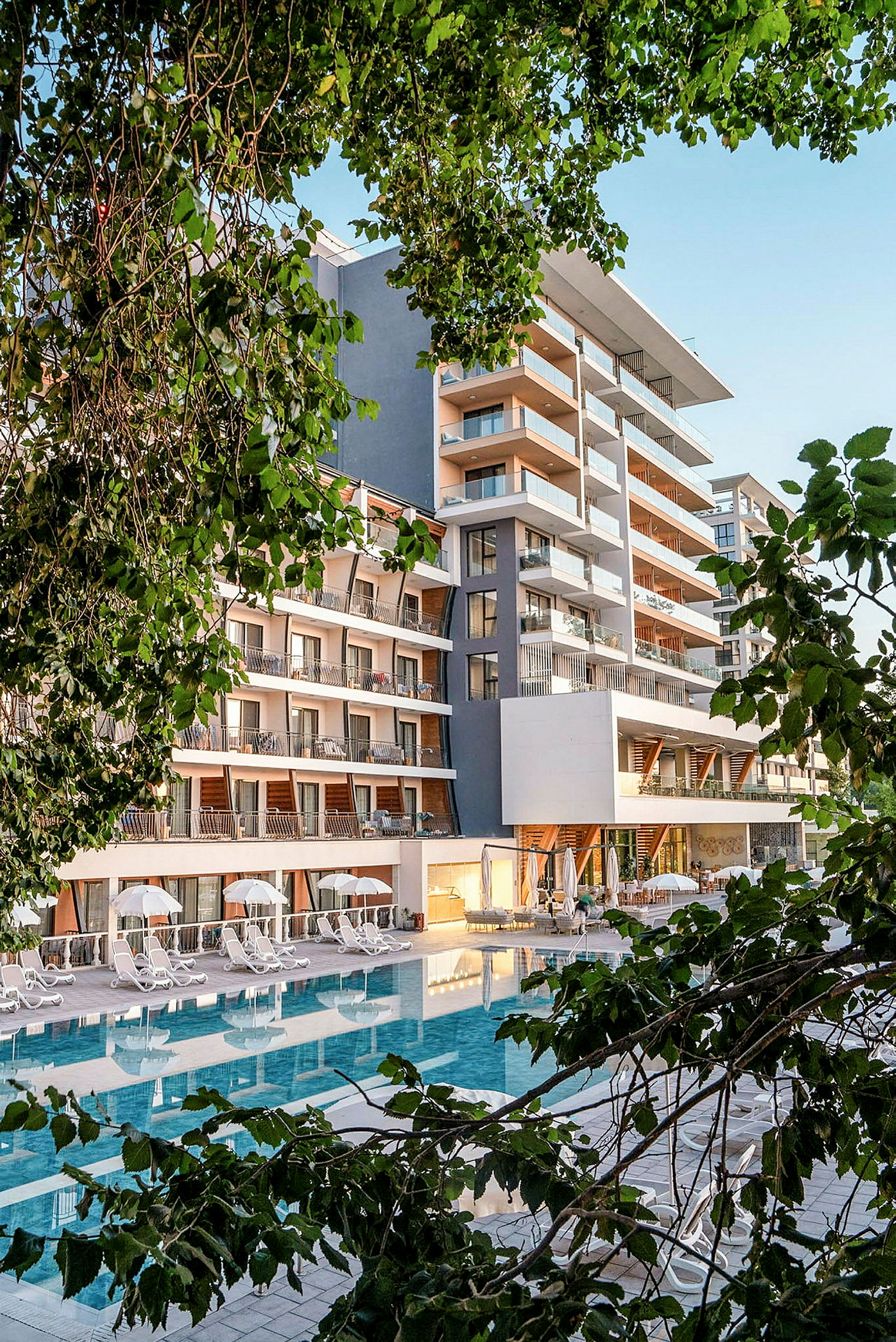 Hotelbygning med pool i forgrunden, omgivet af frodige træer. (AI-genereret tekst)