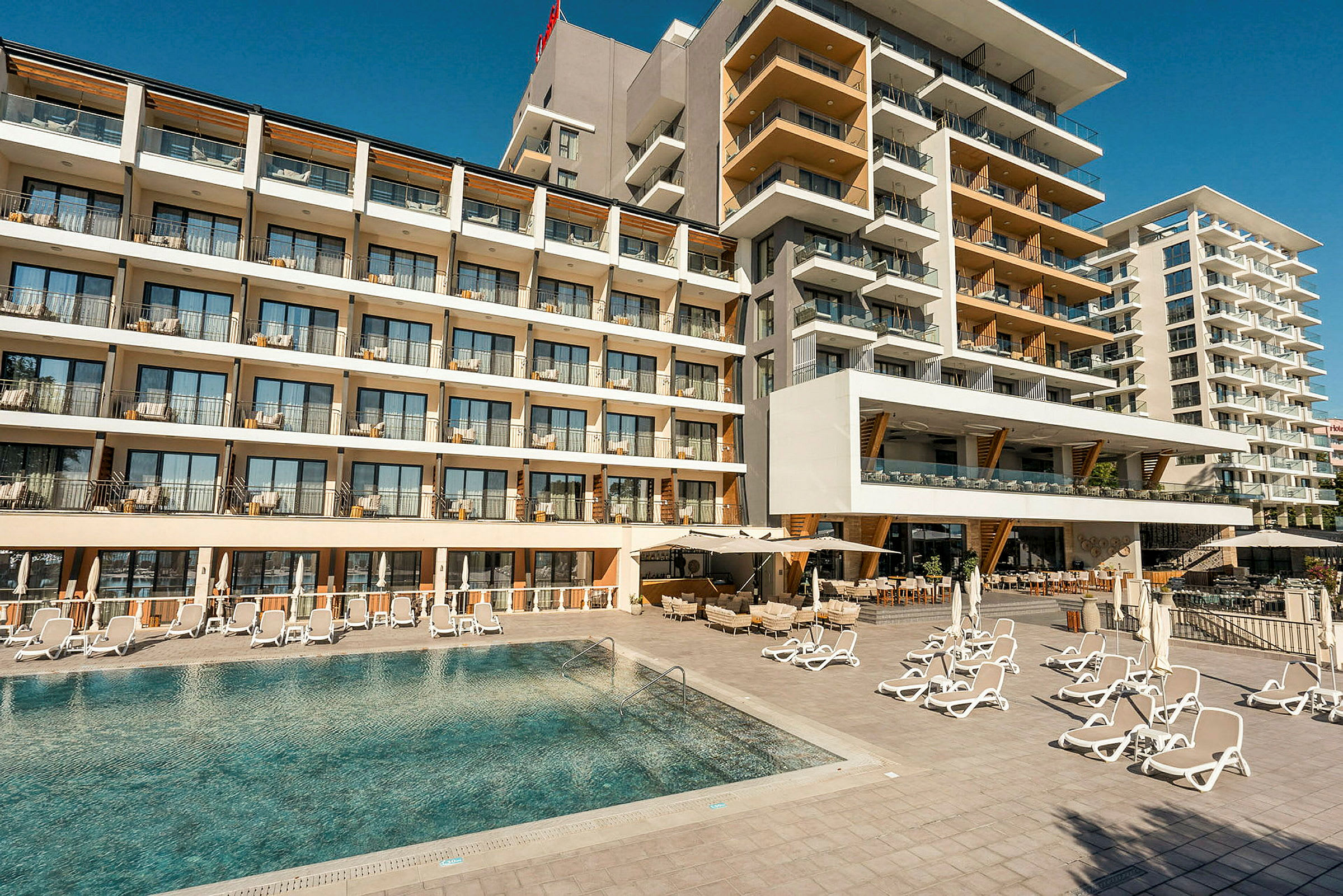 Hotelfacade med flere etager og altaner foran en pool med liggestole. (AI-genereret tekst)