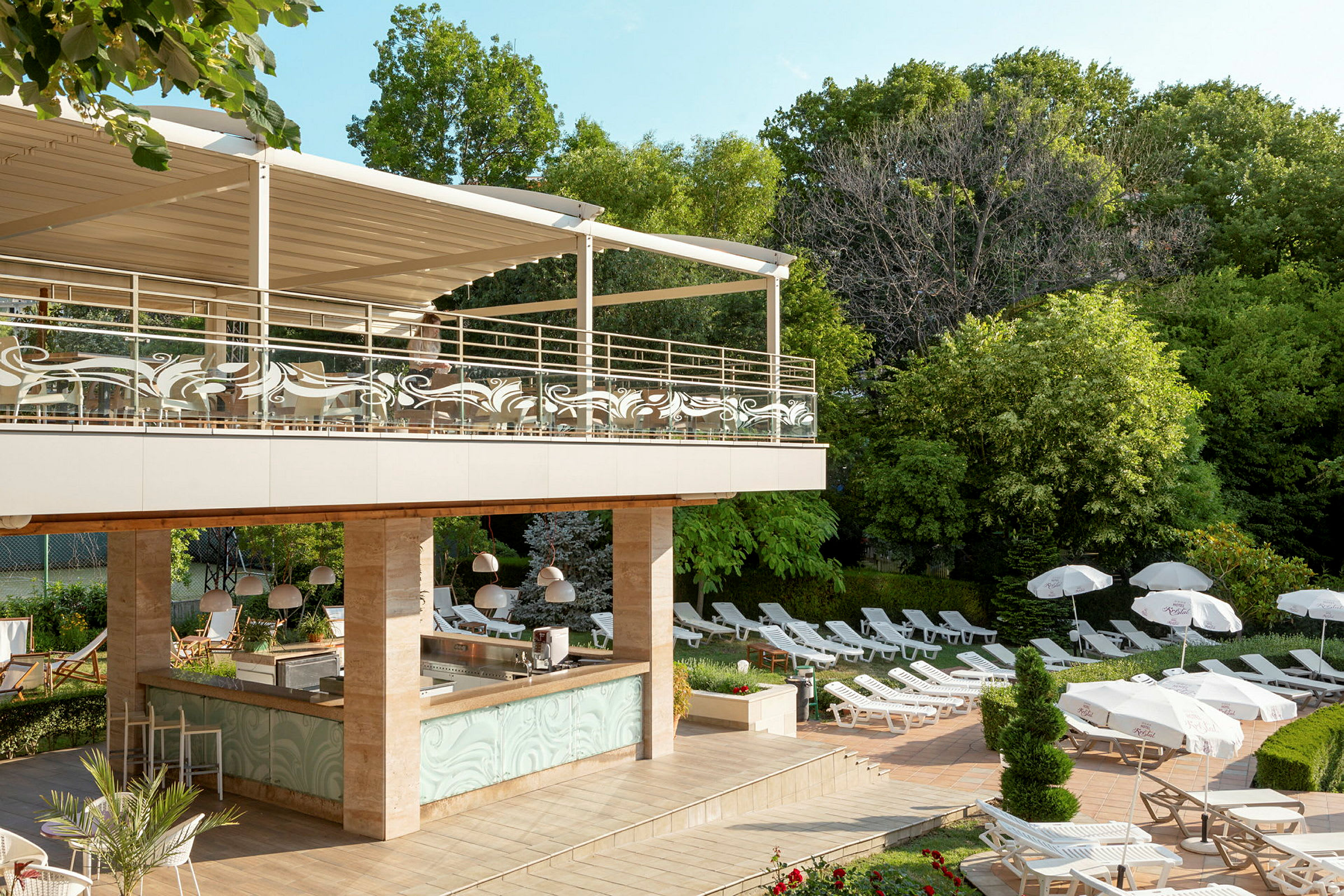 Et udendørs poolområde med liggestole, parasoller og en bar under et overdækket terrasseområde. Området er omgivet af grønne træer. (AI-genereret tekst)