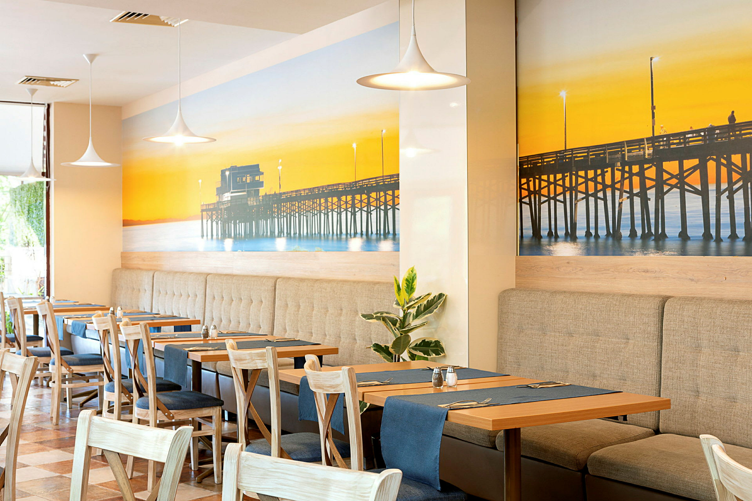 En restaurant med indretning, der inkluderer borde og stole, samt en vægdekoration med en mole ved solnedgang. (AI-genereret tekst)