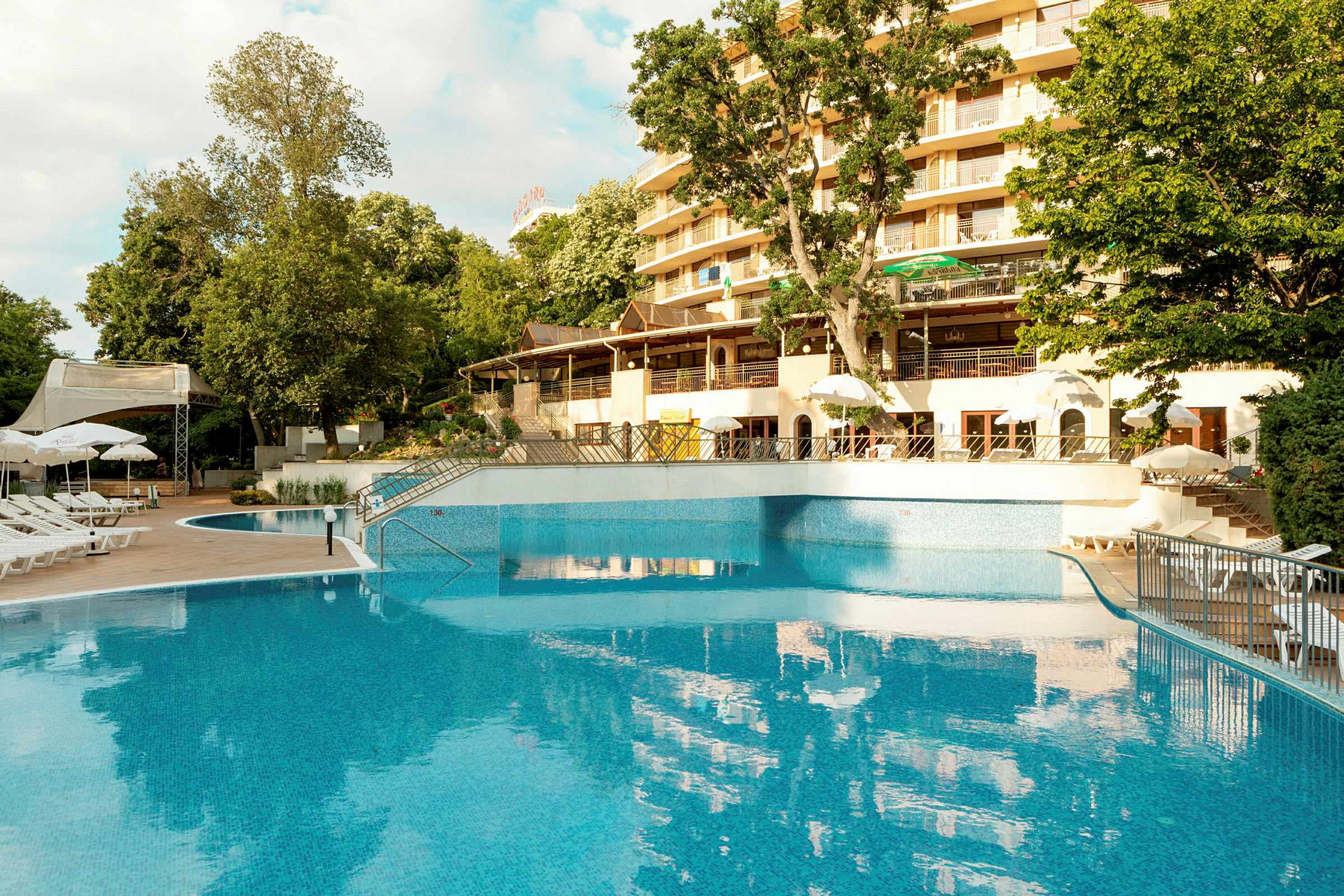 Billede af en pool ved et hotel med liggestole og parasoller samt grønne områder og træer i baggrunden. (AI-genereret tekst)