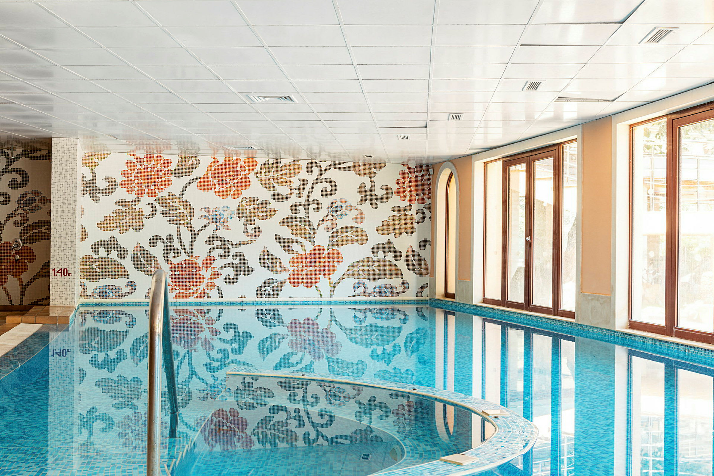 Indendørs pool med blomster-mosaikvæg og store vinduer. (AI-genereret tekst)