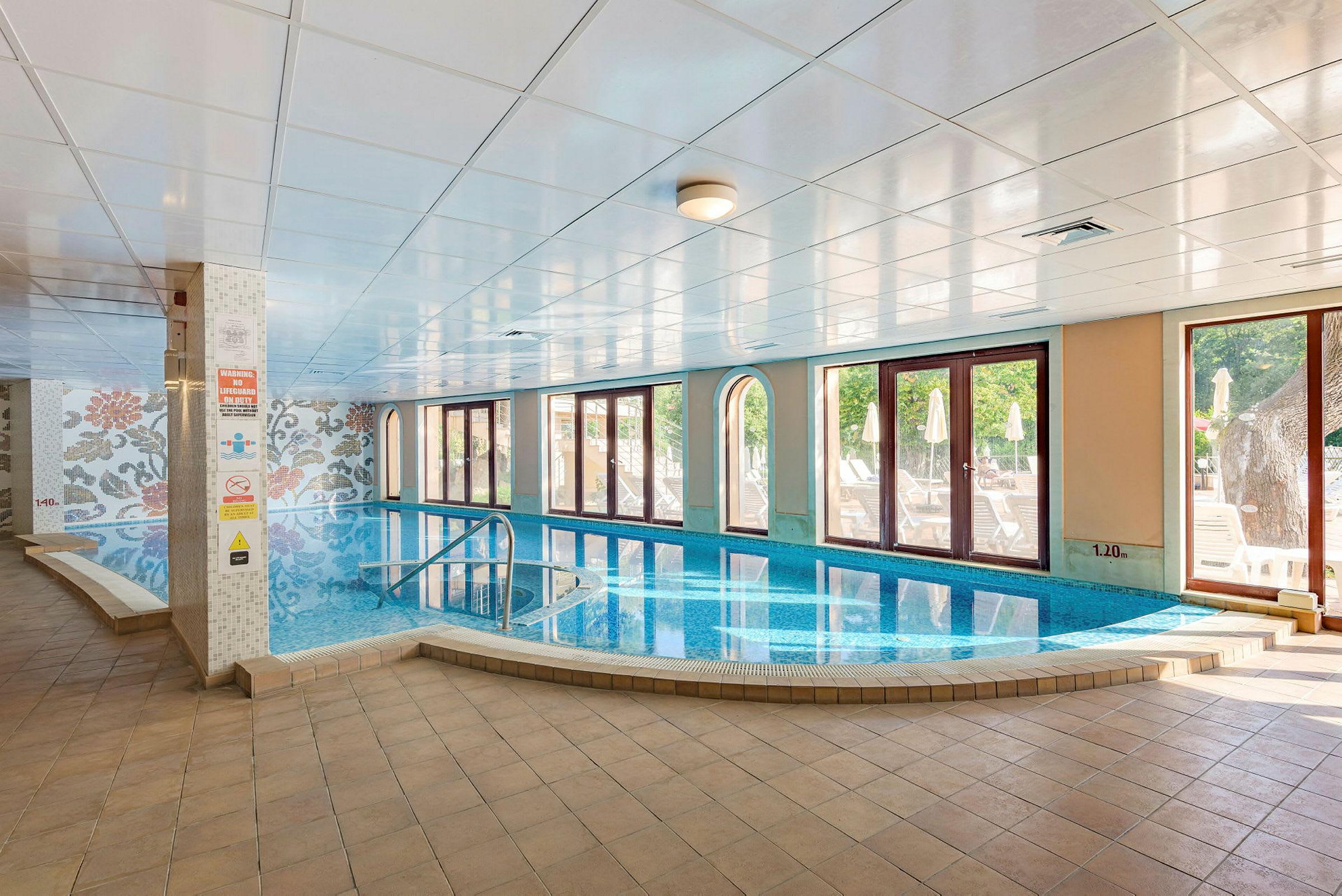 Indendørs pool med store vinduer ud mod en solrig terrasse. (AI-genereret tekst)