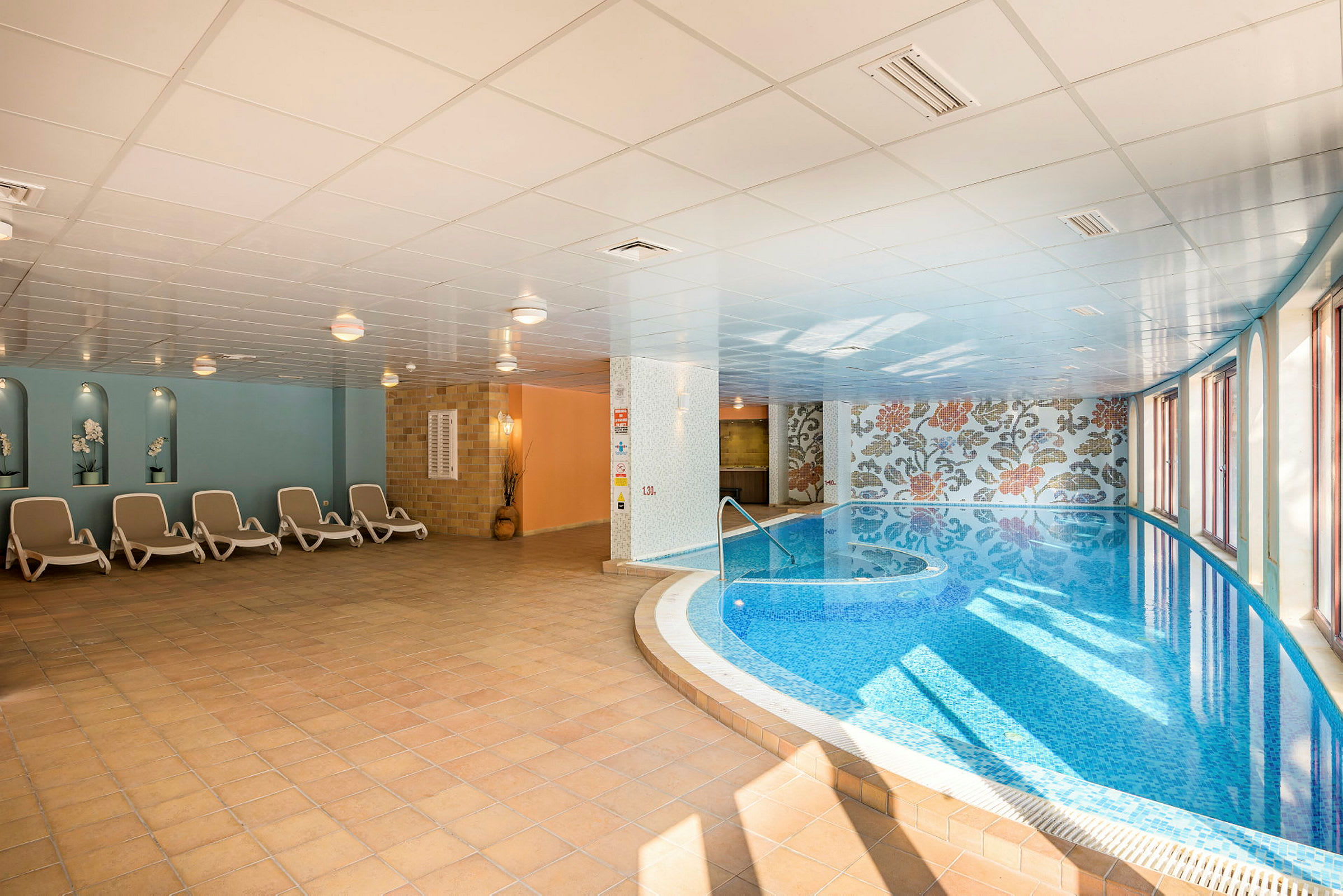 En indendørs pool med blå vand og liggestole ved siden af. (AI-genereret tekst)