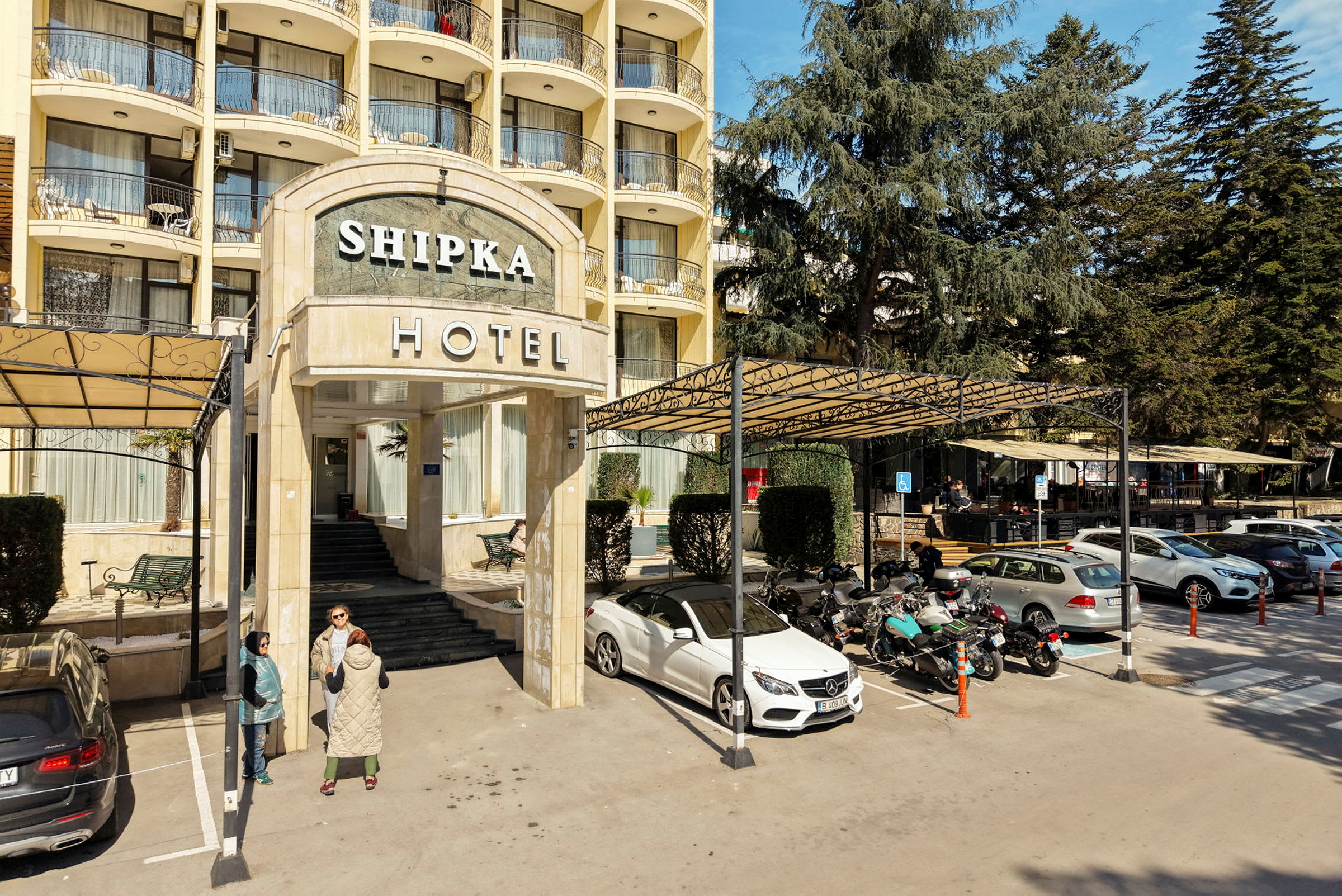 Hotelbygningens indgang med trappe og skilt, omgivet af parkering med biler og motorcykler. (AI-genereret tekst)