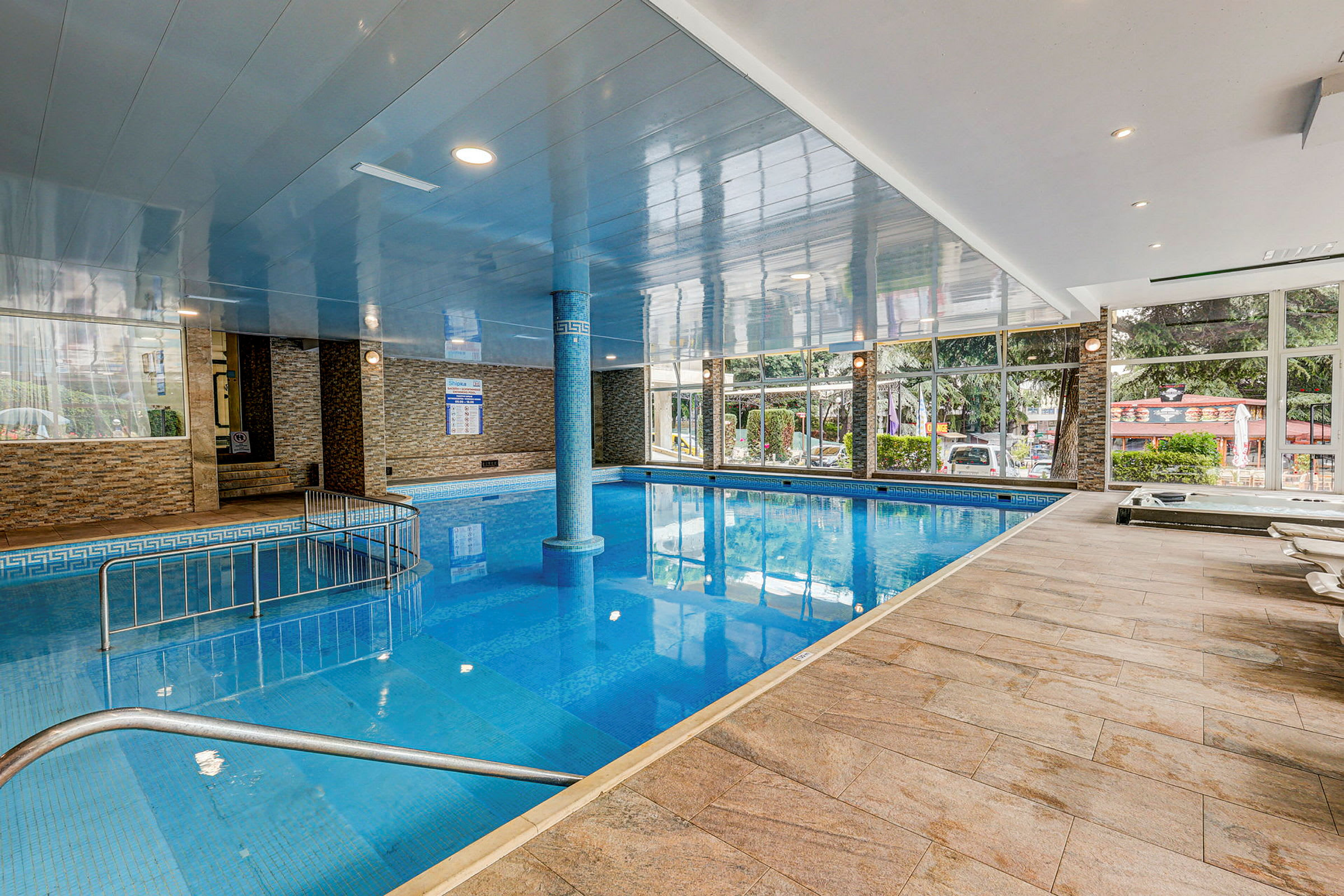En indendørs swimmingpool med flisegulv og glasvægge, der vender ud mod det fri. Der er en søjle midt i poolen samt liggestole langs kanten. (AI-genereret tekst)