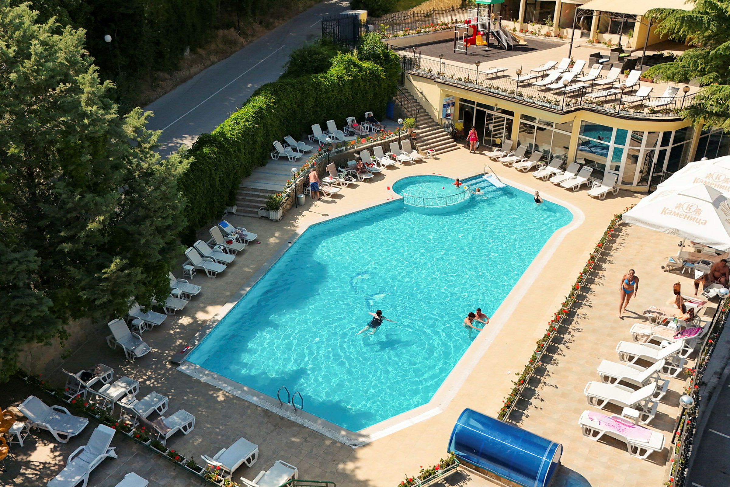 En udendørs swimmingpool omgivet af liggestole og beplantning, med personer der svømmer og slapper af. (AI-genereret tekst)