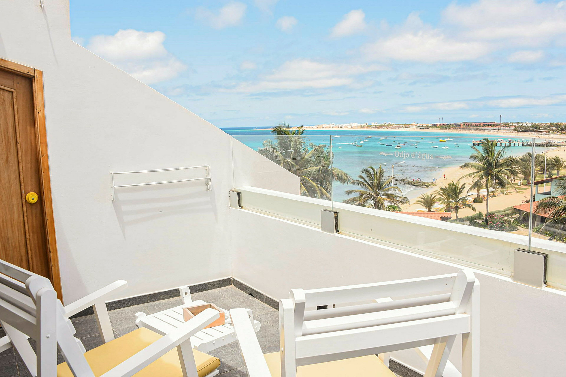 Billedet viser en terrasse med hvide stole og bord samt udsigt over en strand med palmer, hav og små både. (AI-genereret tekst)