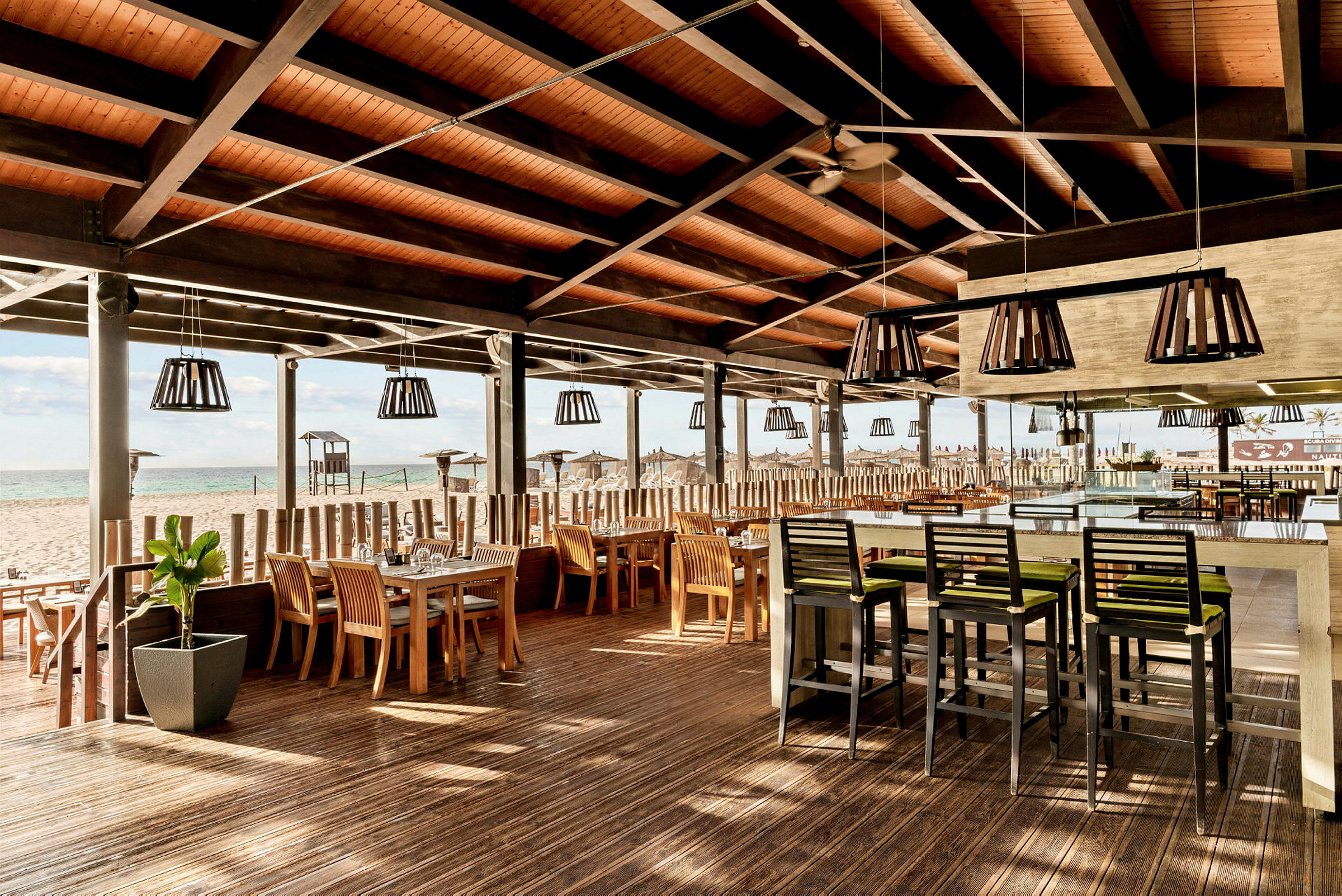 Udendørs restaurant med tag, træmøbler og borde ved en strand. (AI-genereret tekst)
