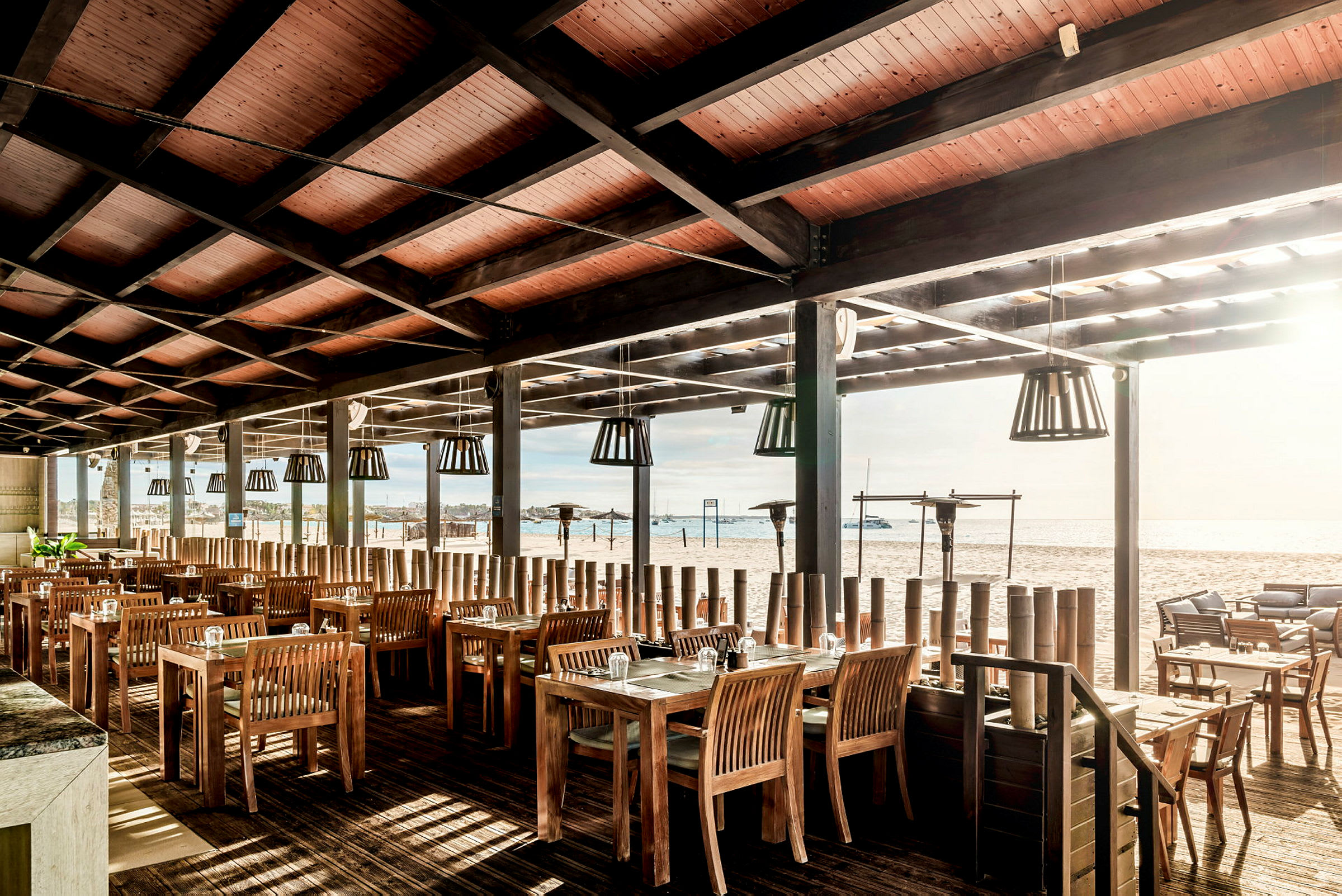 Restaurant med træmøbler og tag, ved stranden med udsigt over vandet. (AI-genereret tekst)