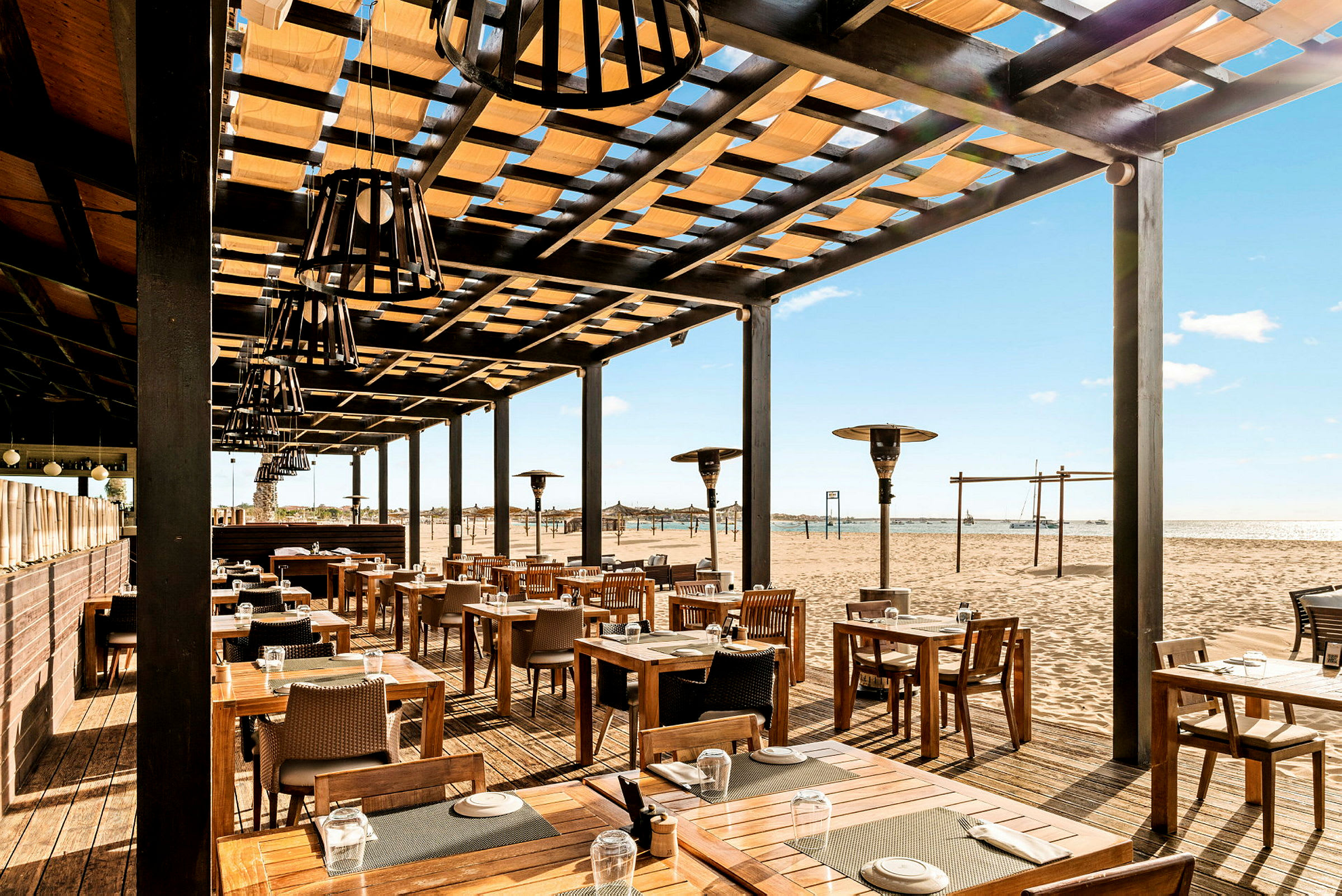 Udendørsrestaurant med træborde og stole, beliggende ved en sandstrand. (AI-genereret tekst)