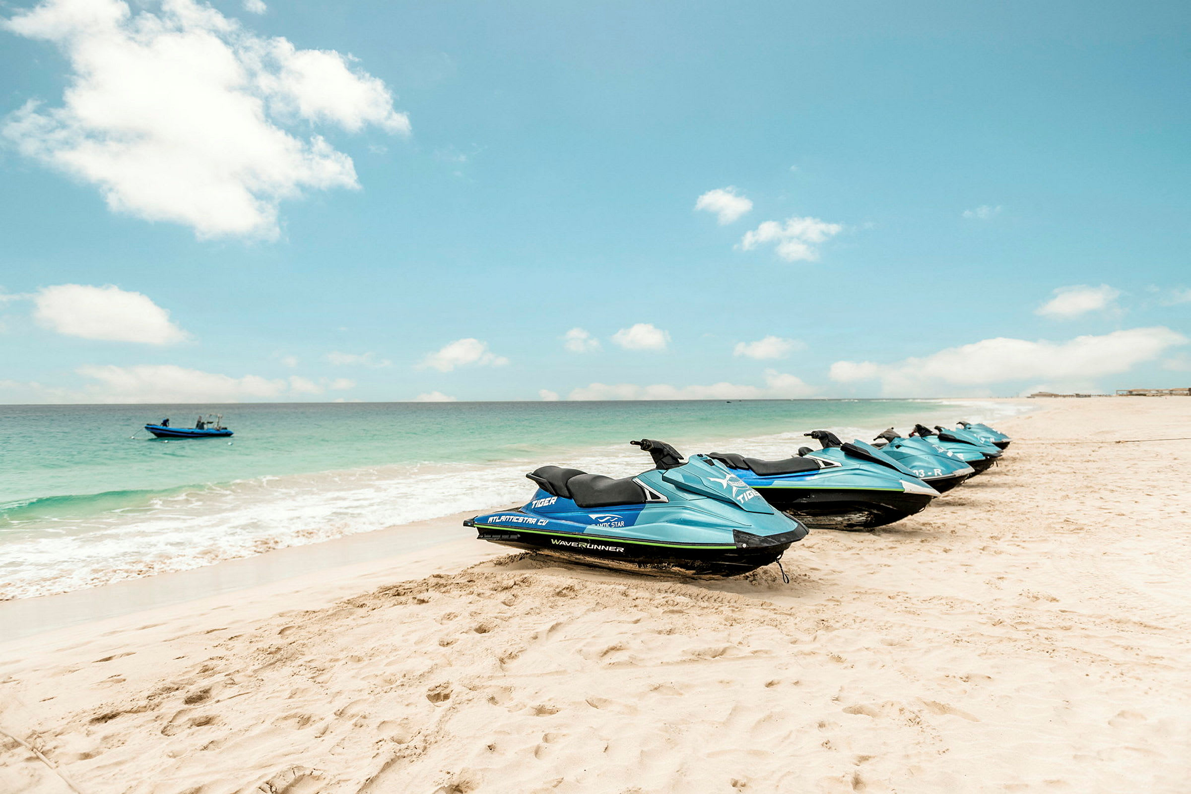 Flere blå jetski står på række på en sandstrand ved havet. (AI-genereret tekst)