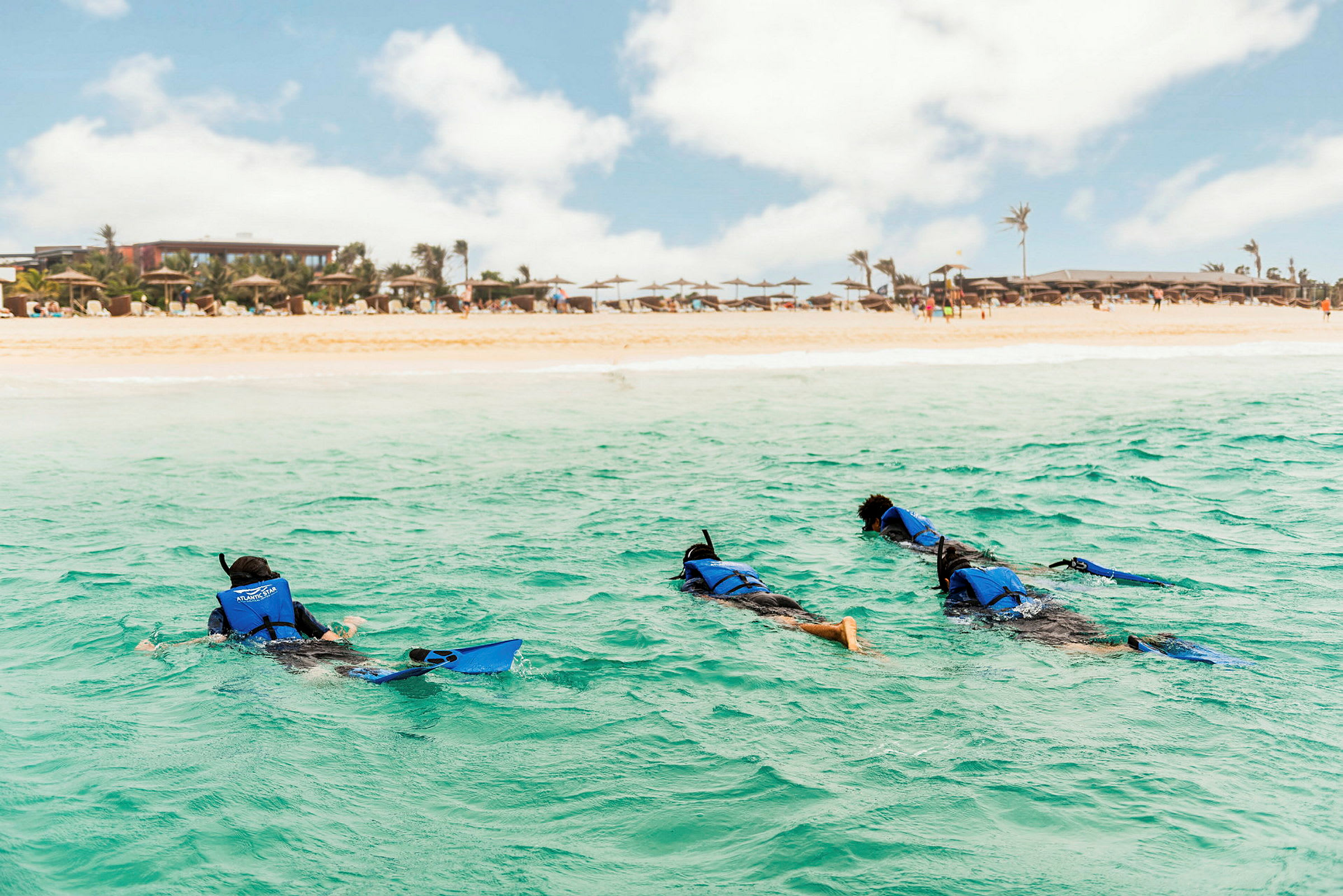 Tre personer snorkler i havet nær en sandstrand med parasoller og bygninger i baggrunden. (AI-genereret tekst)
