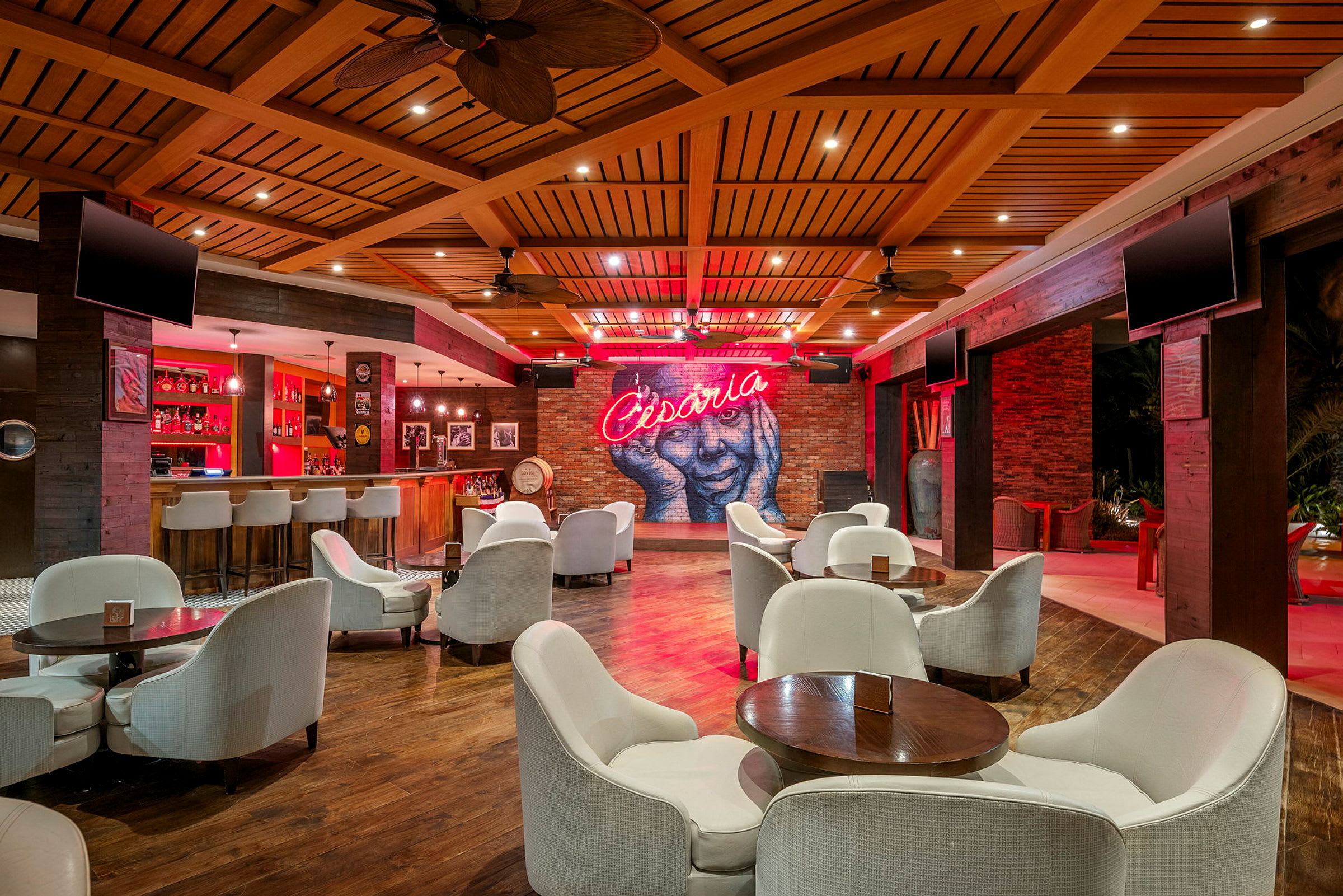 En elegant lounge med hvide stole og borde, en bar i baggrunden og en væg med en neontekst, der siger 'Cesaria'. (AI-genereret tekst)