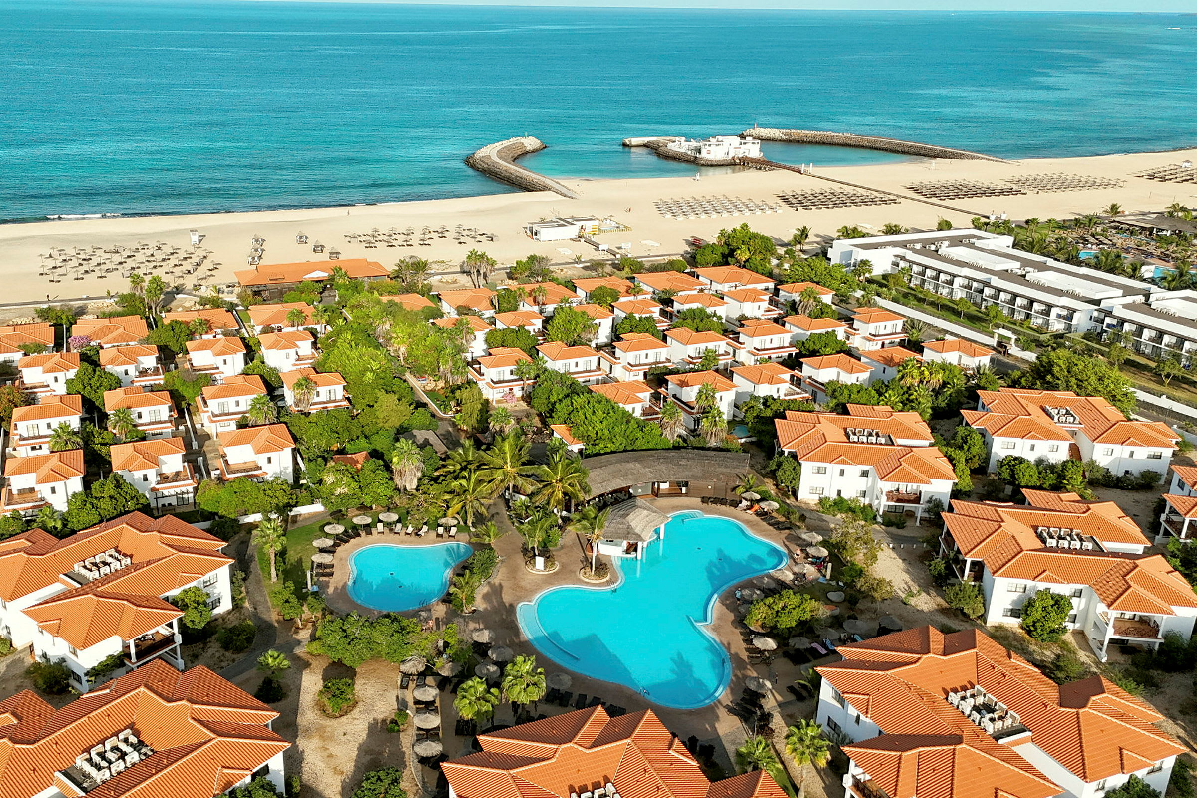 Luftfoto af et ferieområde med hvide huse med orange tage, swimmingpool og en sandstrand ved havet. (AI-genereret tekst)