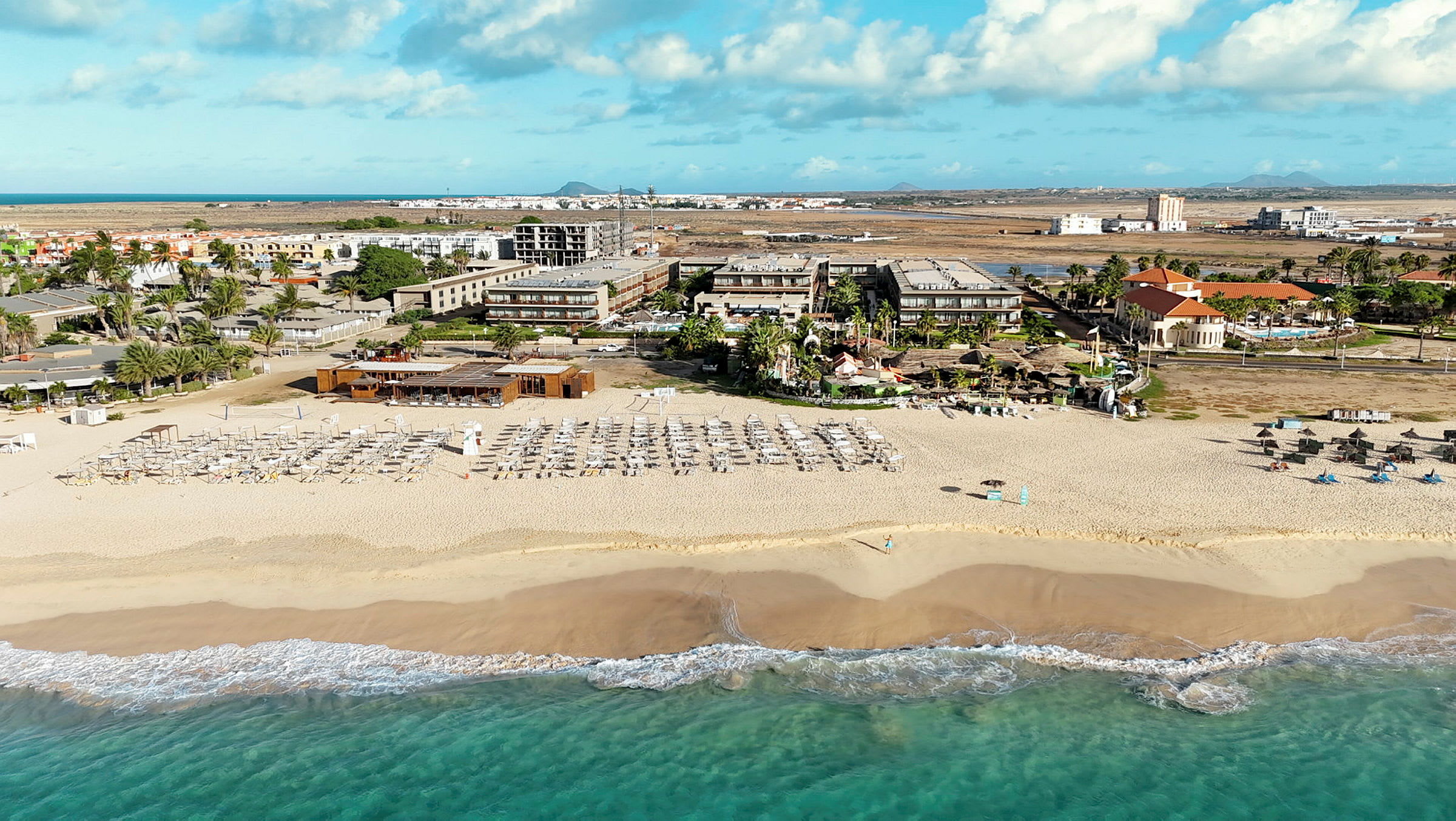 Billede af en sandstrand med solvogne og parasoller, hotelbygninger i baggrunden. (AI-genereret tekst)
