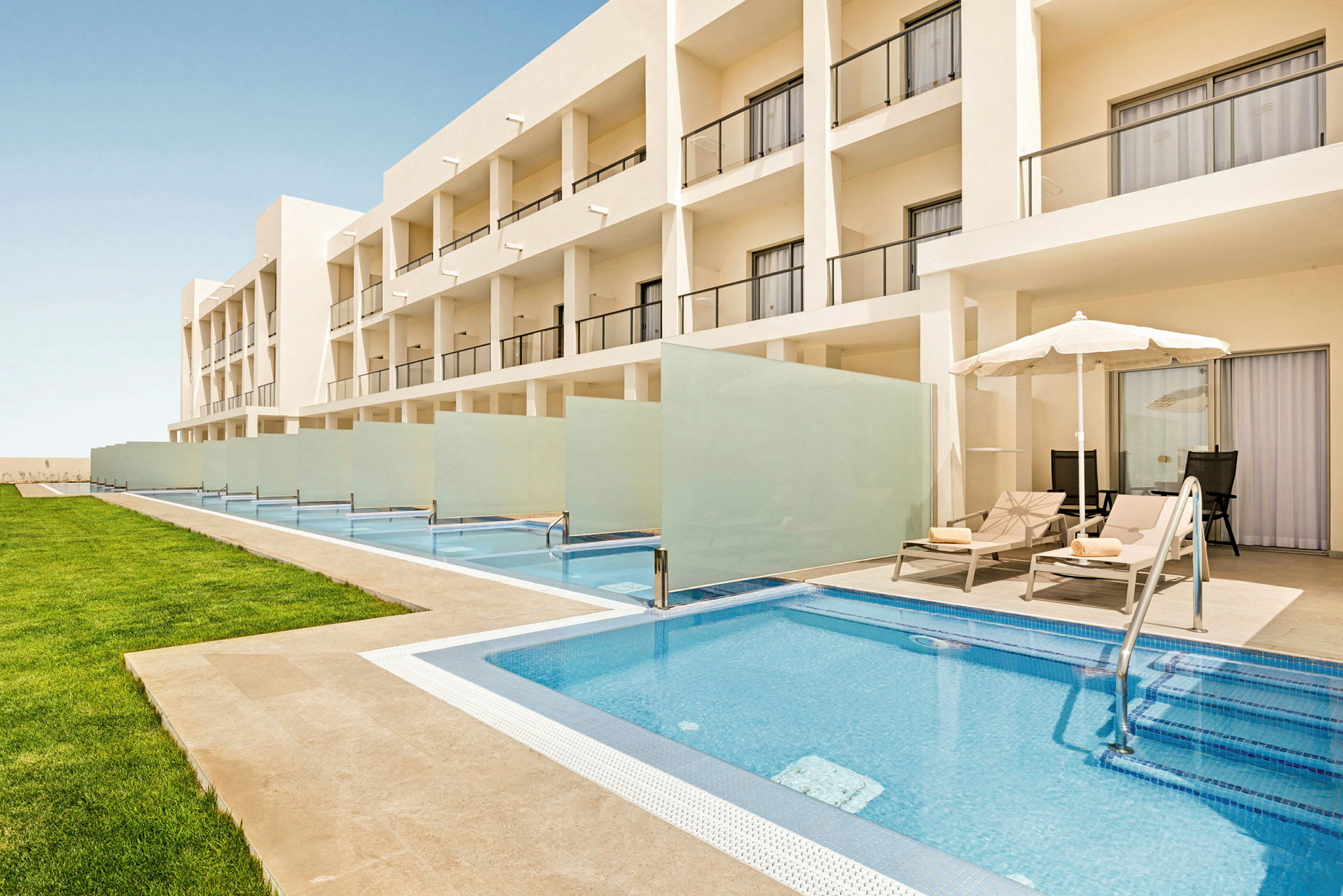 Billede af en moderne hotelbygning med private pools foran hvert værelse og liggestole under parasoller ved siden af poolsene. (AI-genereret tekst)