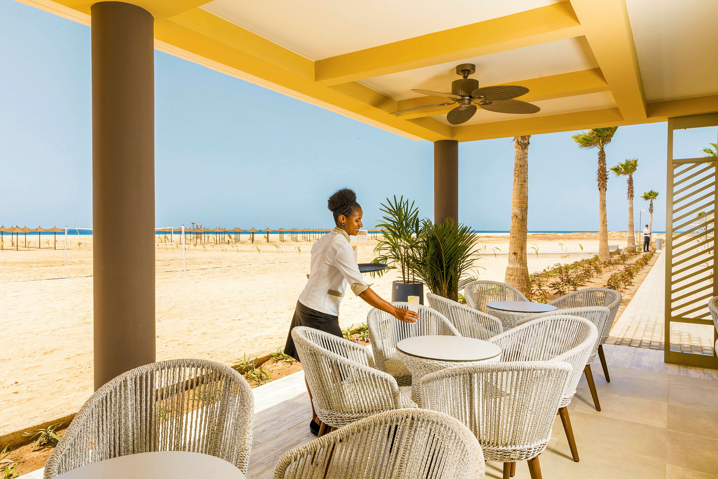 En udendørs terrasse med moderne møbler og udsigt over en sandstrand og havet i baggrunden. (AI-genereret tekst)