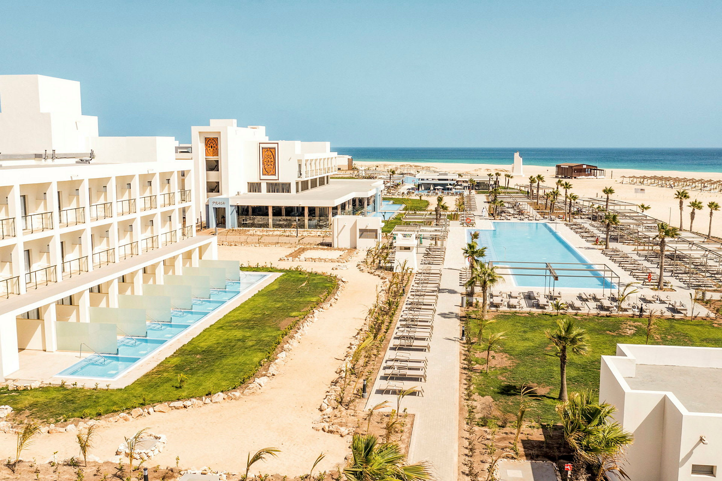 Et moderne hotel med hvide bygninger, der omgiver flere pools og frodige områder, beliggende tæt på en sandstrand ved havet. (AI-genereret tekst)
