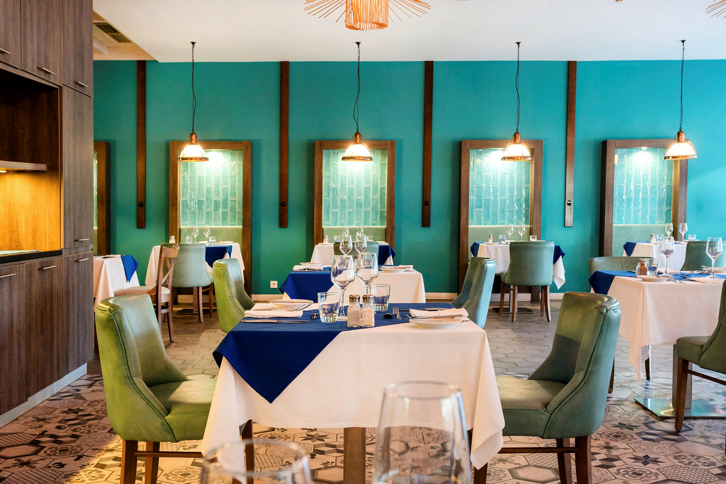 Et elegant restaurantinteriør med dækkede borde og grønne stole, omgivet af turkise vægge og dekorative hængelamper. (AI-genereret tekst)