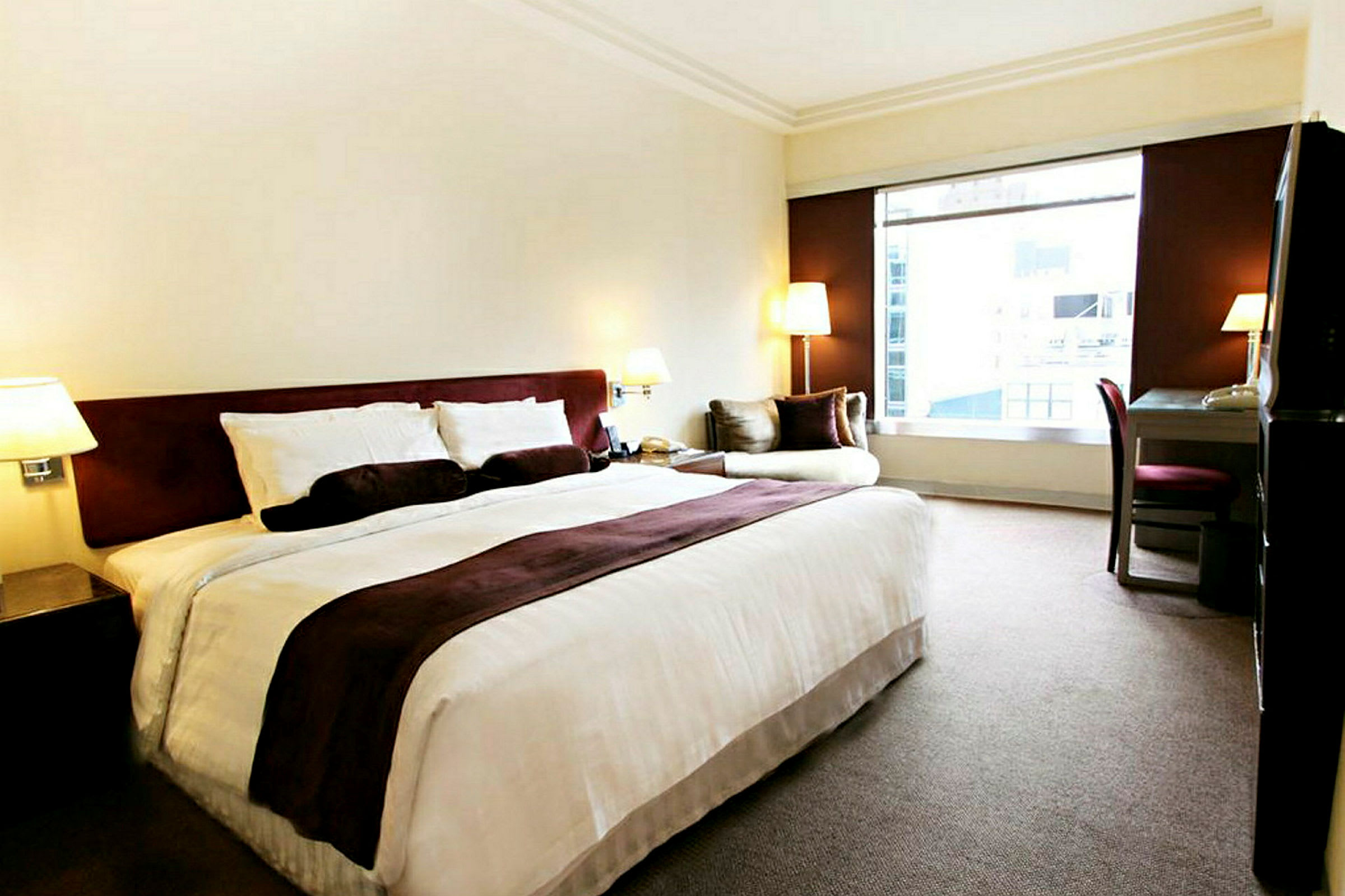 Hotelværelse med en stor seng, et skrivebord og en lænestol ved siden af et stort vindue. (AI-genereret tekst)