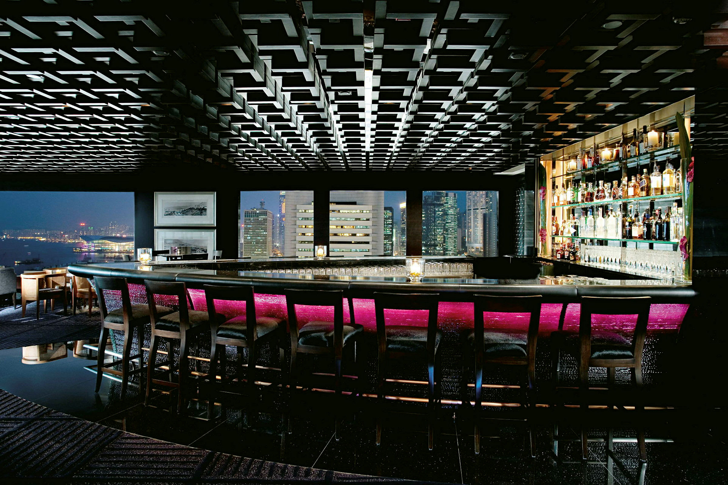 Billede af en luksuriøs bar med høje rygstole ved bardisken. Baren har stemningsfuld belysning med en neonrosa nuance og store vinduer, der viser udsigten over byens skyline. (AI-genereret tekst)