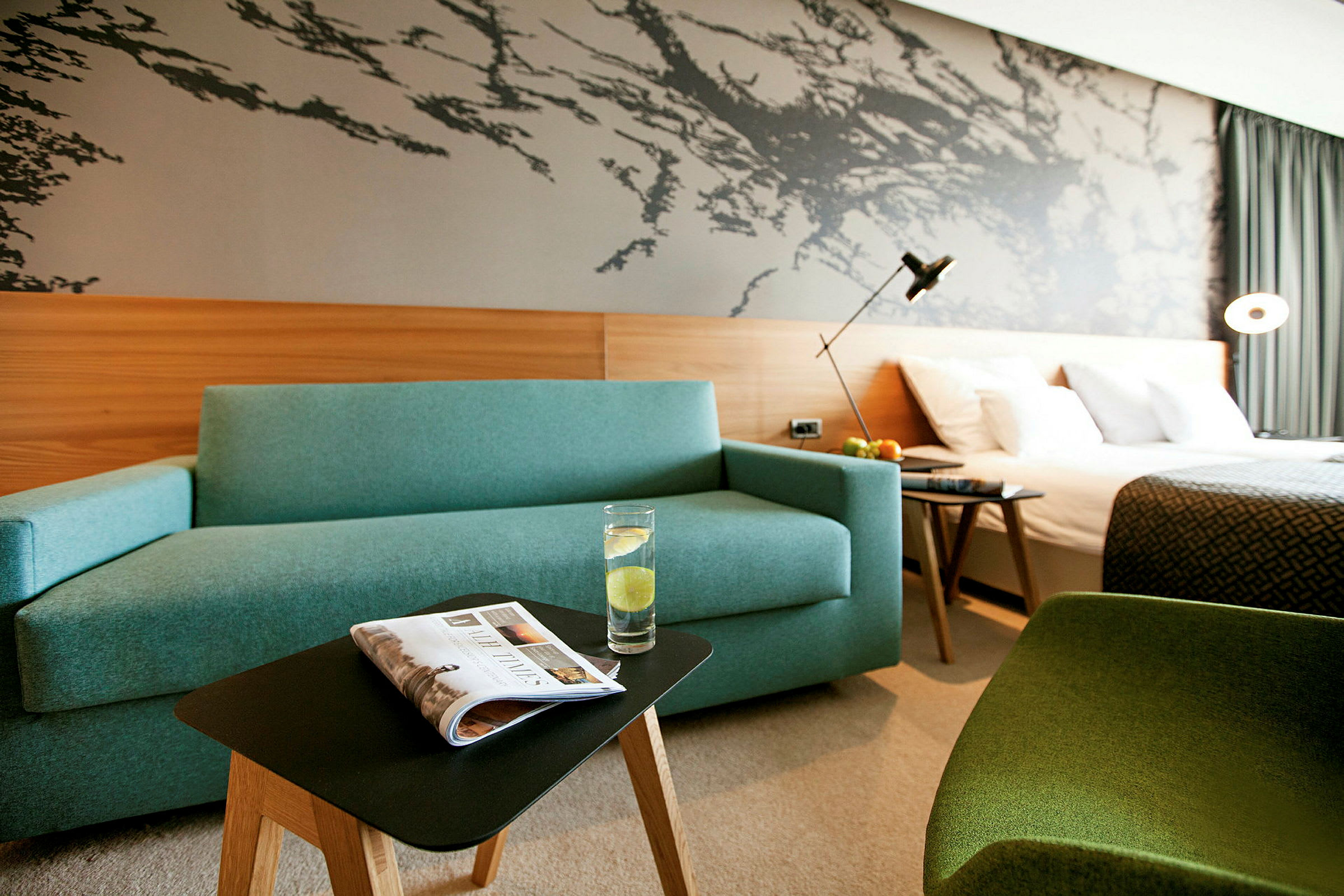 Hotelværelse med en blå sofa, et lille bord med et glas vand, en seng og en dekorativ væg. (AI-genereret tekst)