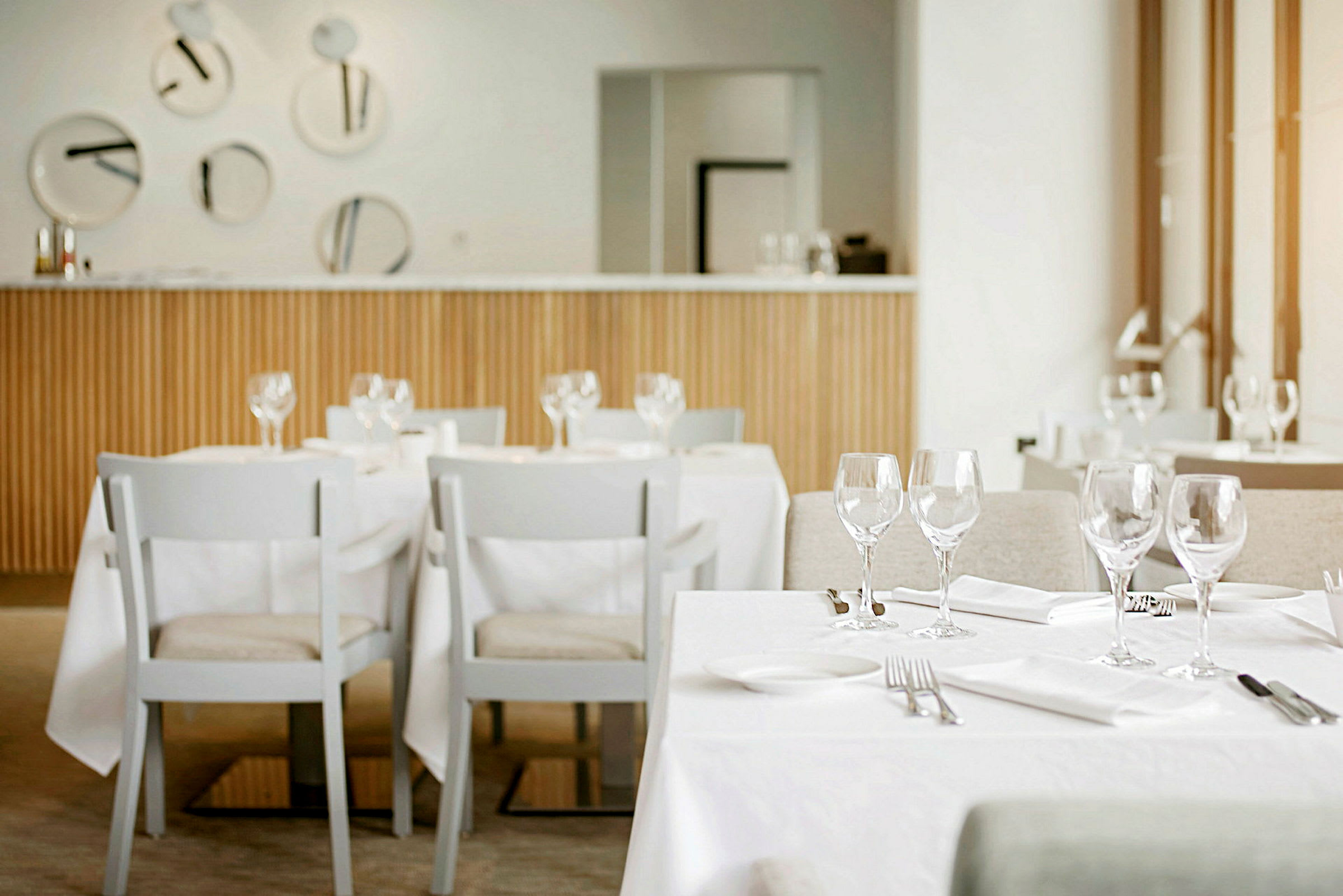En moderne og lys restaurantindretning med opdækkede borde og hvide stole. På bordene er der glas, bestik og servietter. (AI-genereret tekst)
