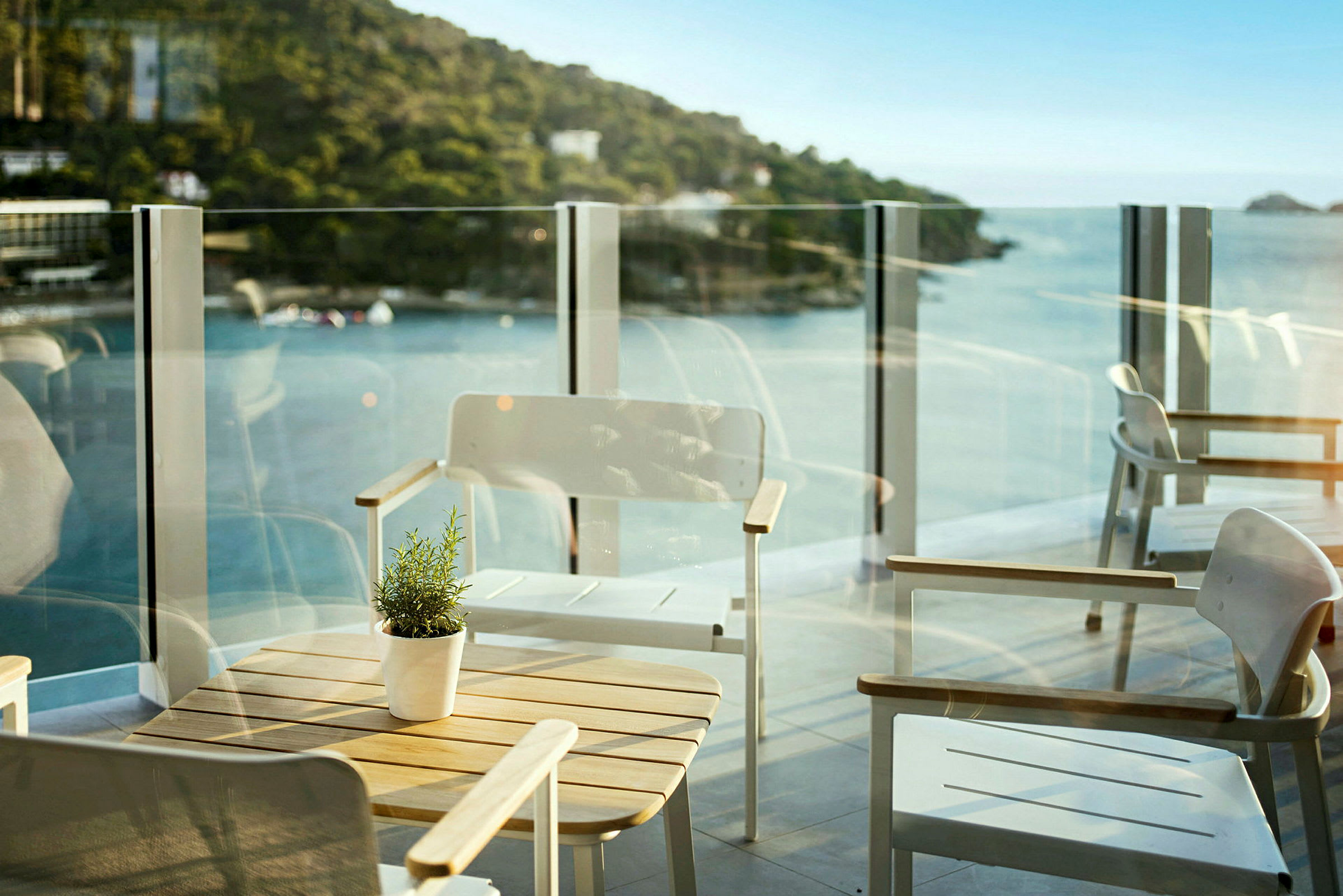 En balkon med moderne møbler og en fantastisk udsigt over havet og skovklædte bakker. (AI-genereret tekst)
