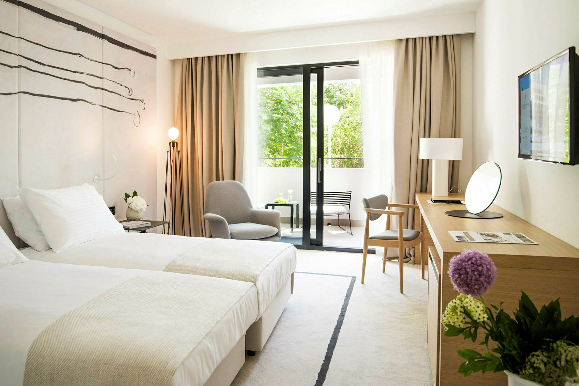 Et moderne hotelværelse med to senge, en lænestol, et skrivebord med et spejl og et TV på væggen. Der er store vinduesdøre, der fører ud til en udendørs altan med udsigt over grønne områder. (AI-genereret tekst)
