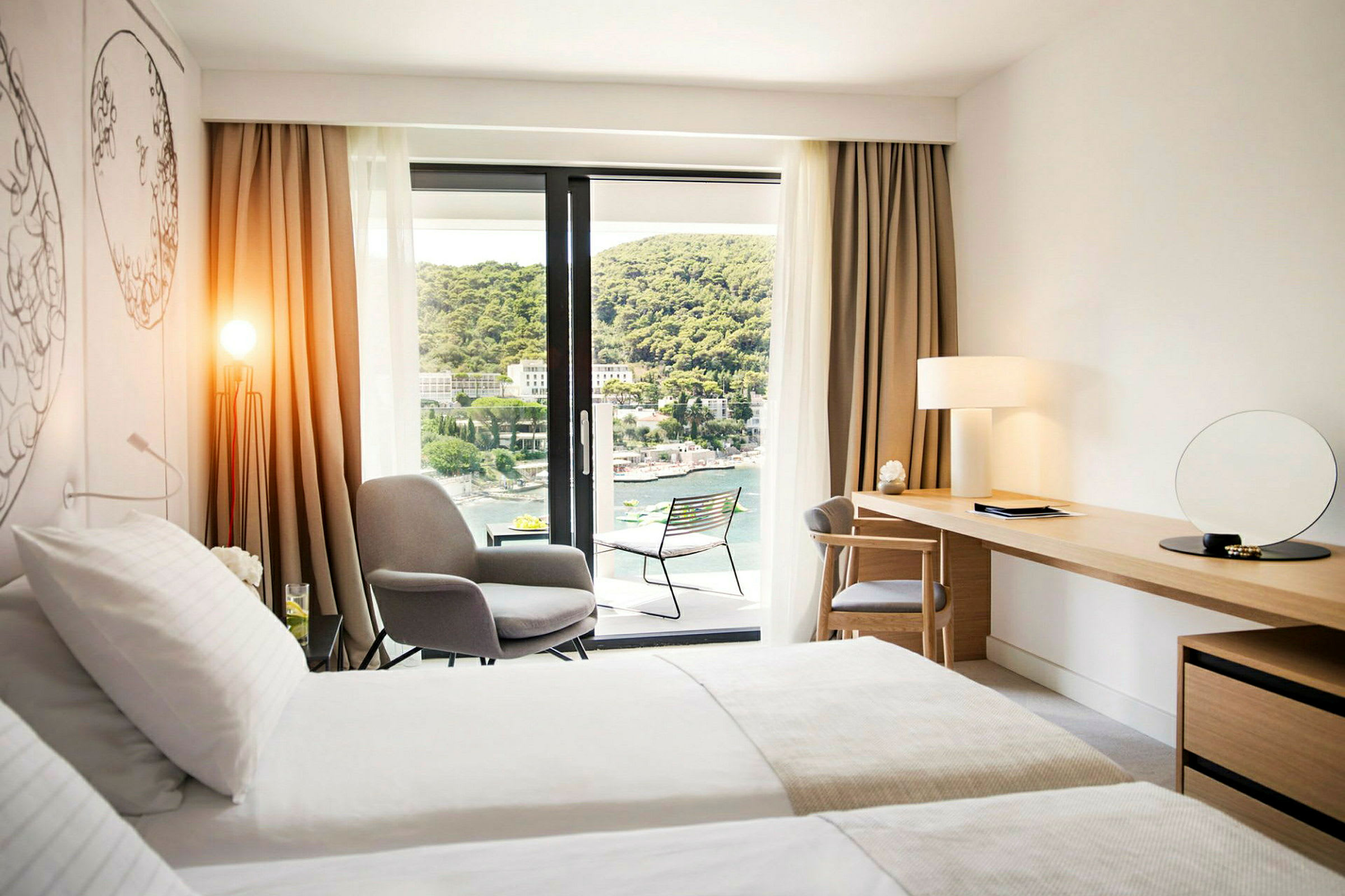 Moderne hotelværelse med dobbeltseng, siddeområde, skrivebord og balkon med udsigt over en frodig skråning og vand. (AI-genereret tekst)