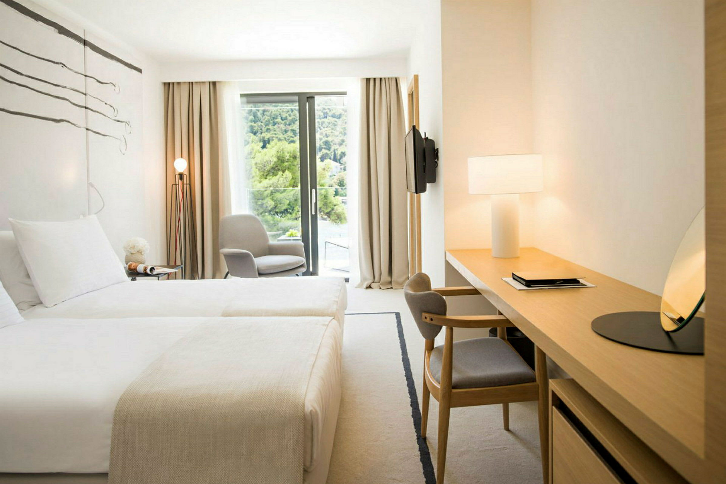 Et lyst hotelværelse med to senge, et skrivebordshjørne, en stol ved et vindue og udsigt over grønt. (AI-genereret tekst)
