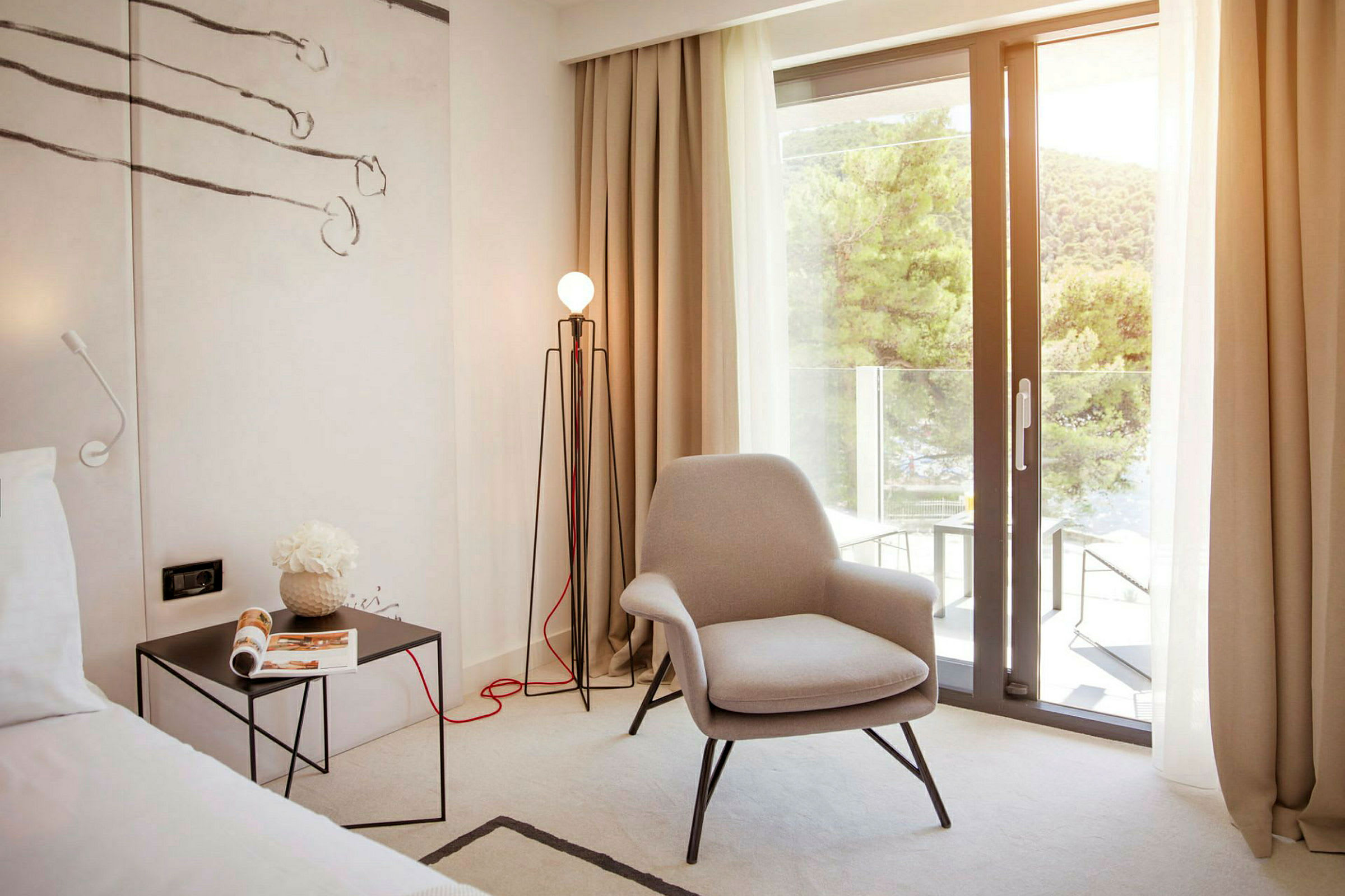 Et hotelværelse med en komfortabel lænestol, et sidebord med et magasin og en gulvlampe. En stor glasdør åbner ud til en altan med udsigt over frodige træer. (AI-genereret tekst)