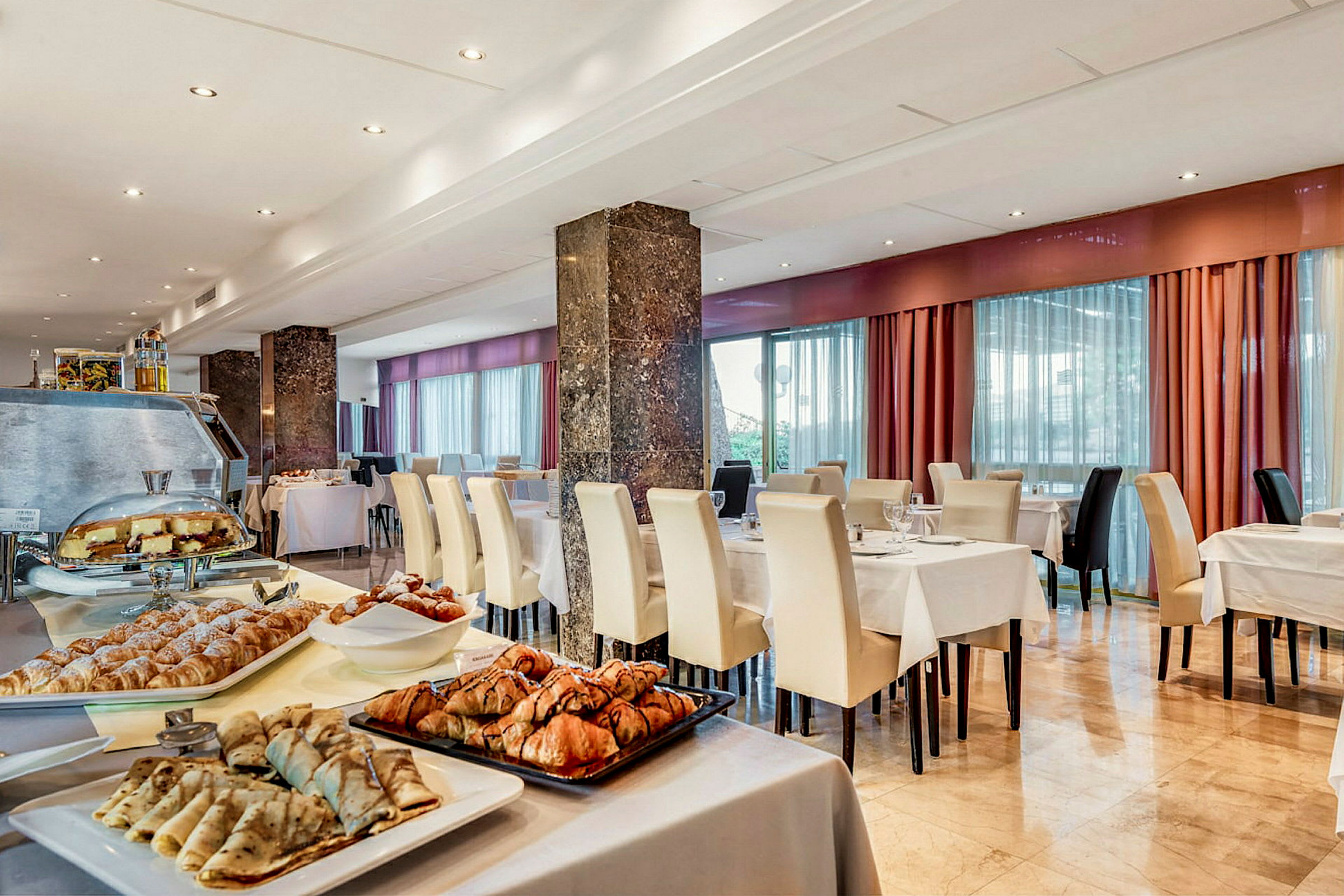 En hotelrestaurant med dækkede borde og hvide stole samt et morgenbuffetbord med forskellige bagværk og pandekager. (AI-genereret tekst)