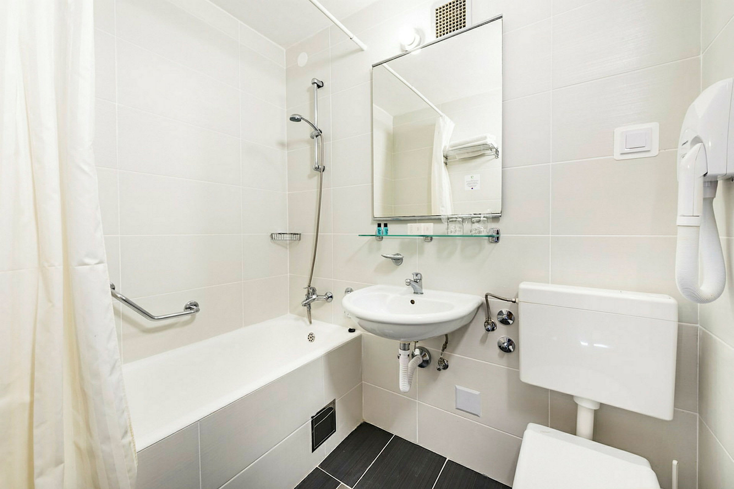 Et moderne badeværelse med badekar, håndvask, toilet og spejl. På væggen er der en hårtørrer og der findes kroge til håndklæder. (AI-genereret tekst)