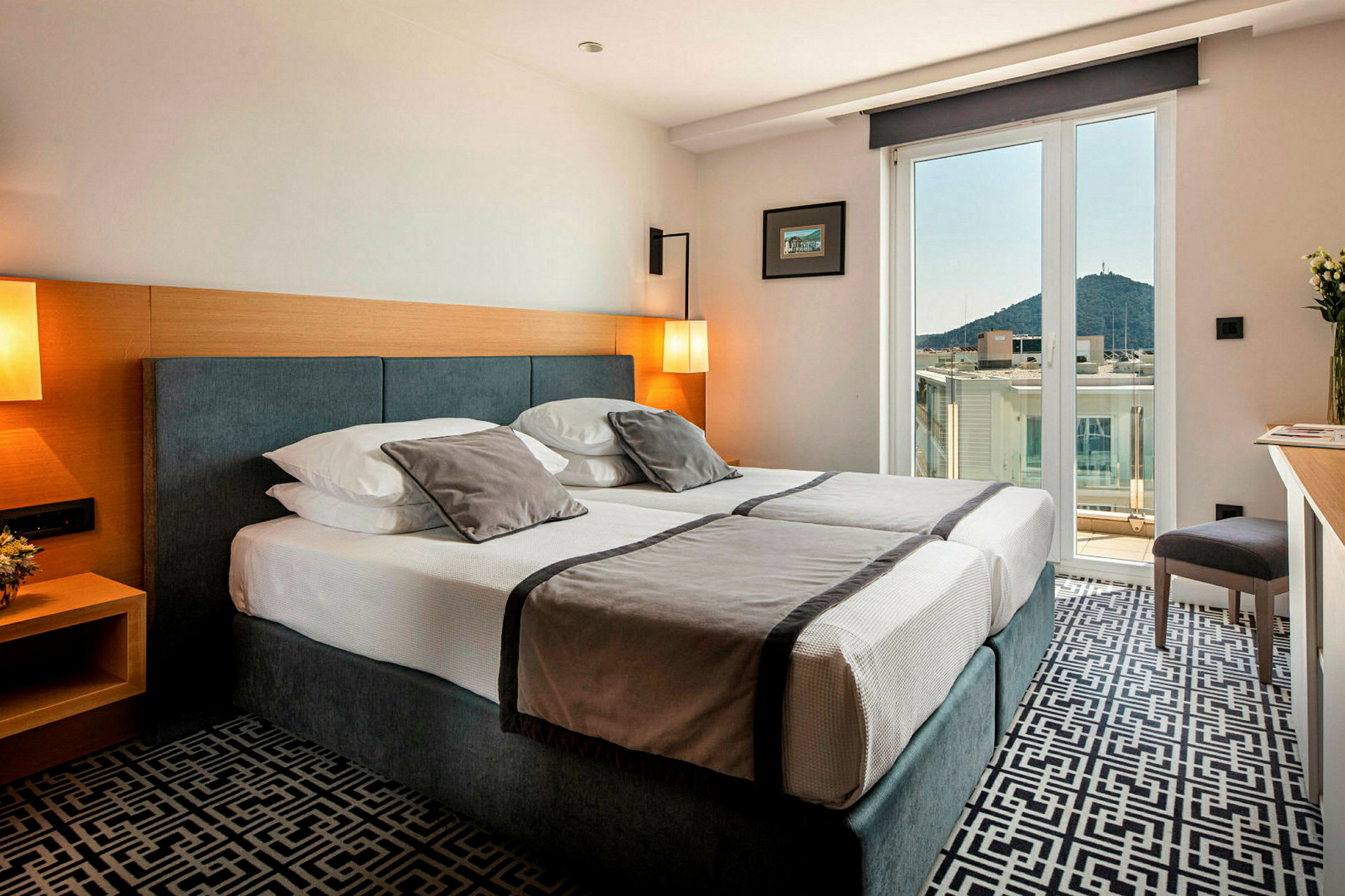 Hotelværelse med dobbeltseng, lys indretning og balkon med udsigt over by og bjerge. (AI-genereret tekst)