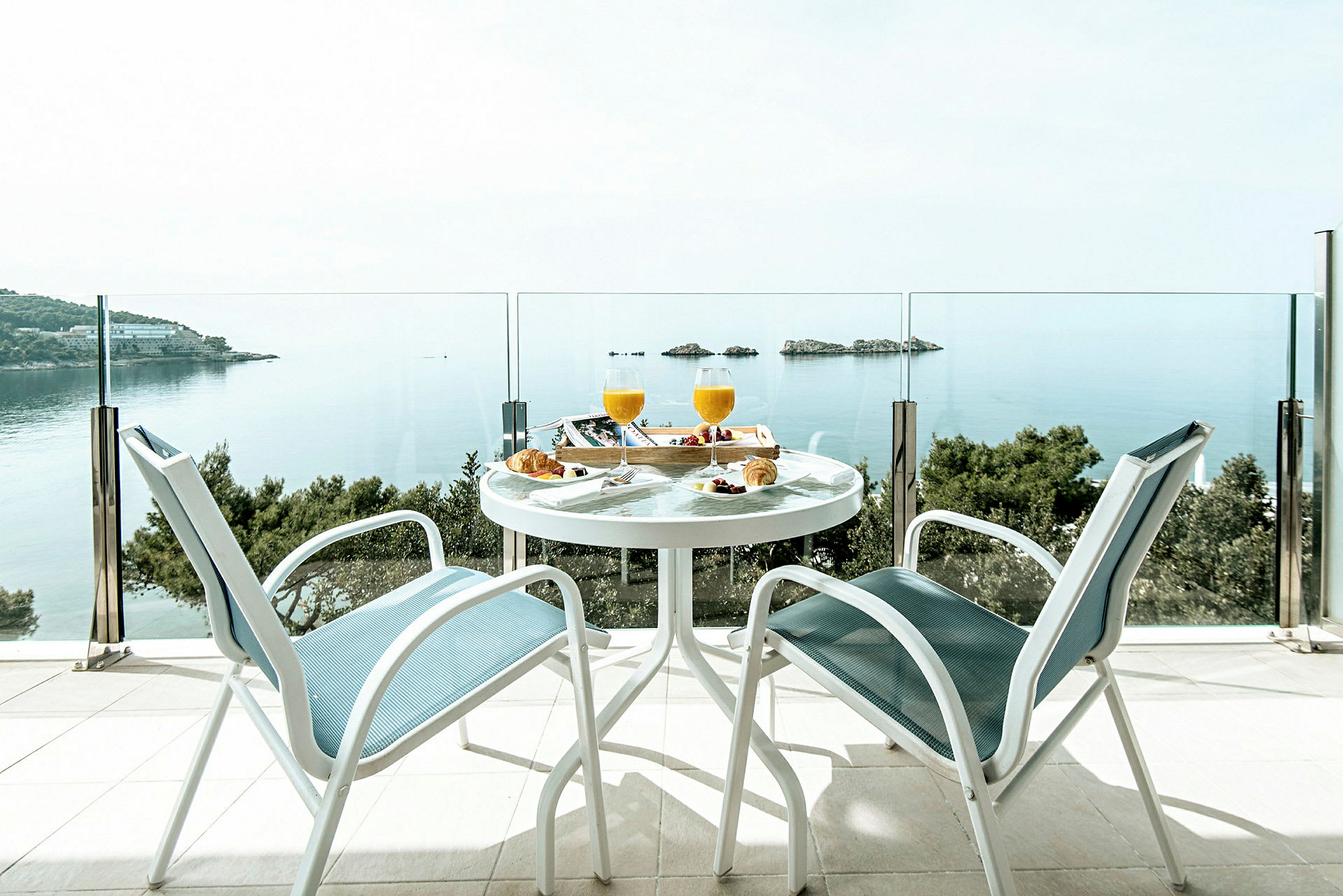 To stole og et bord med morgenmad er placeret på en terrasse med udsigt over havet og nogle små øer. (AI-genereret tekst)