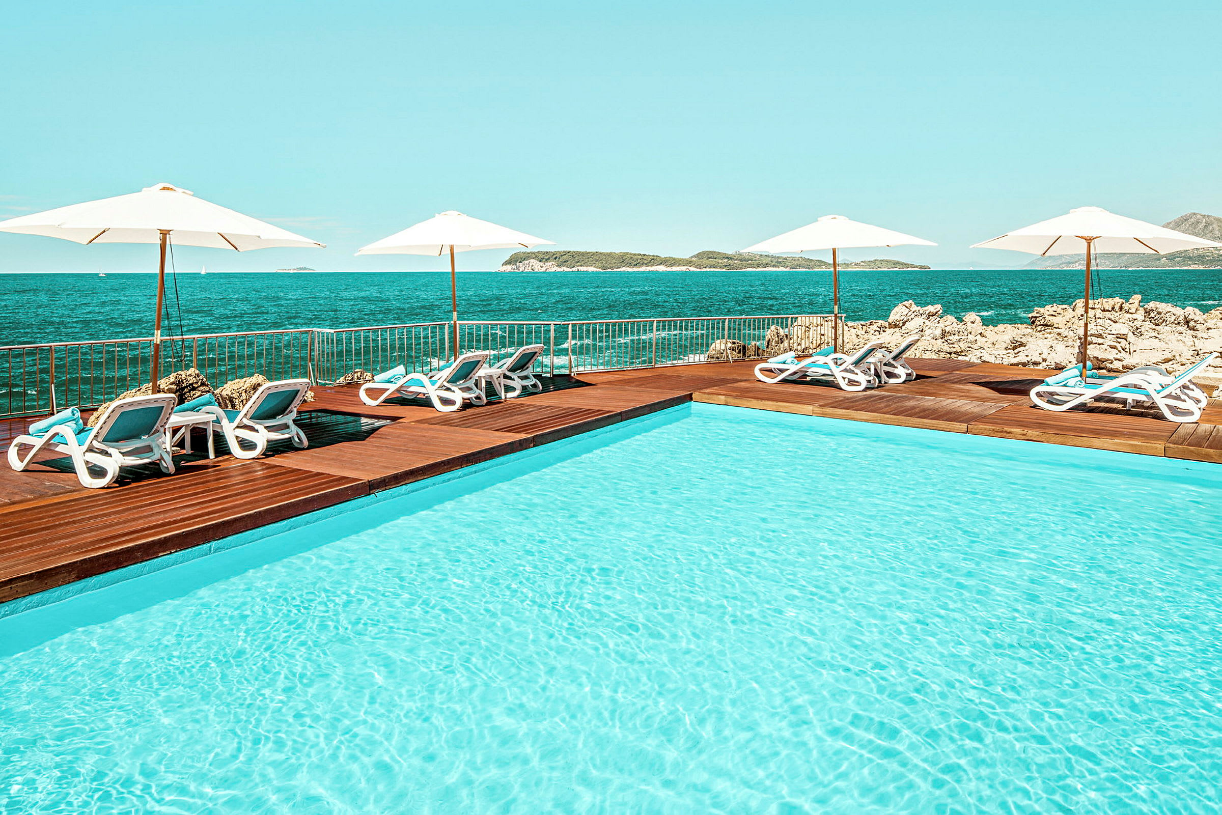 En udendørs pool ved havet med liggestole og parasoller arrangeret ved poolens træterrasse. (AI-genereret tekst)