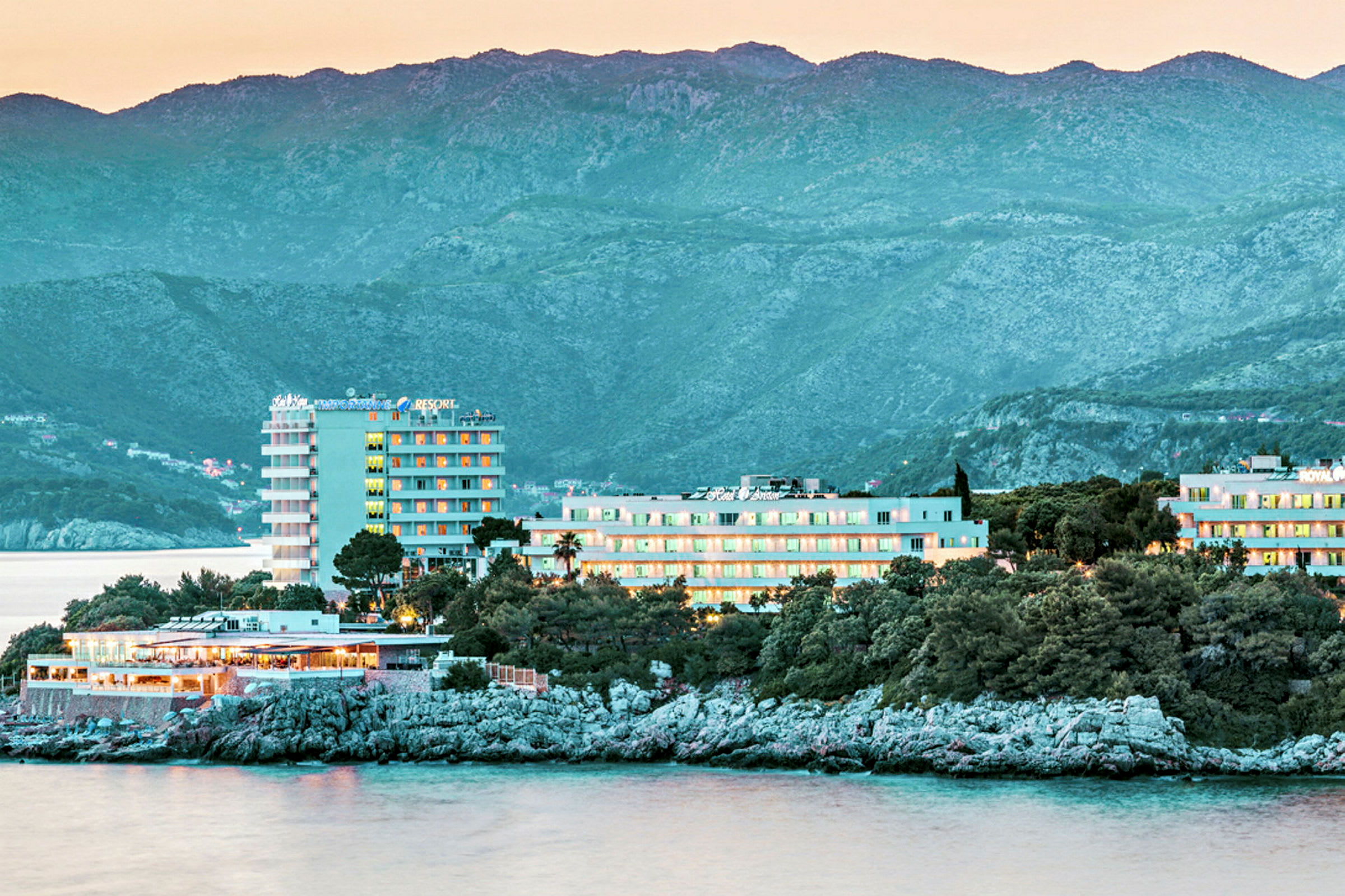 Et kystnært hotel med udsigt over havet, omgivet af træer og grønt, med bjerge i baggrunden. (AI-genereret tekst)