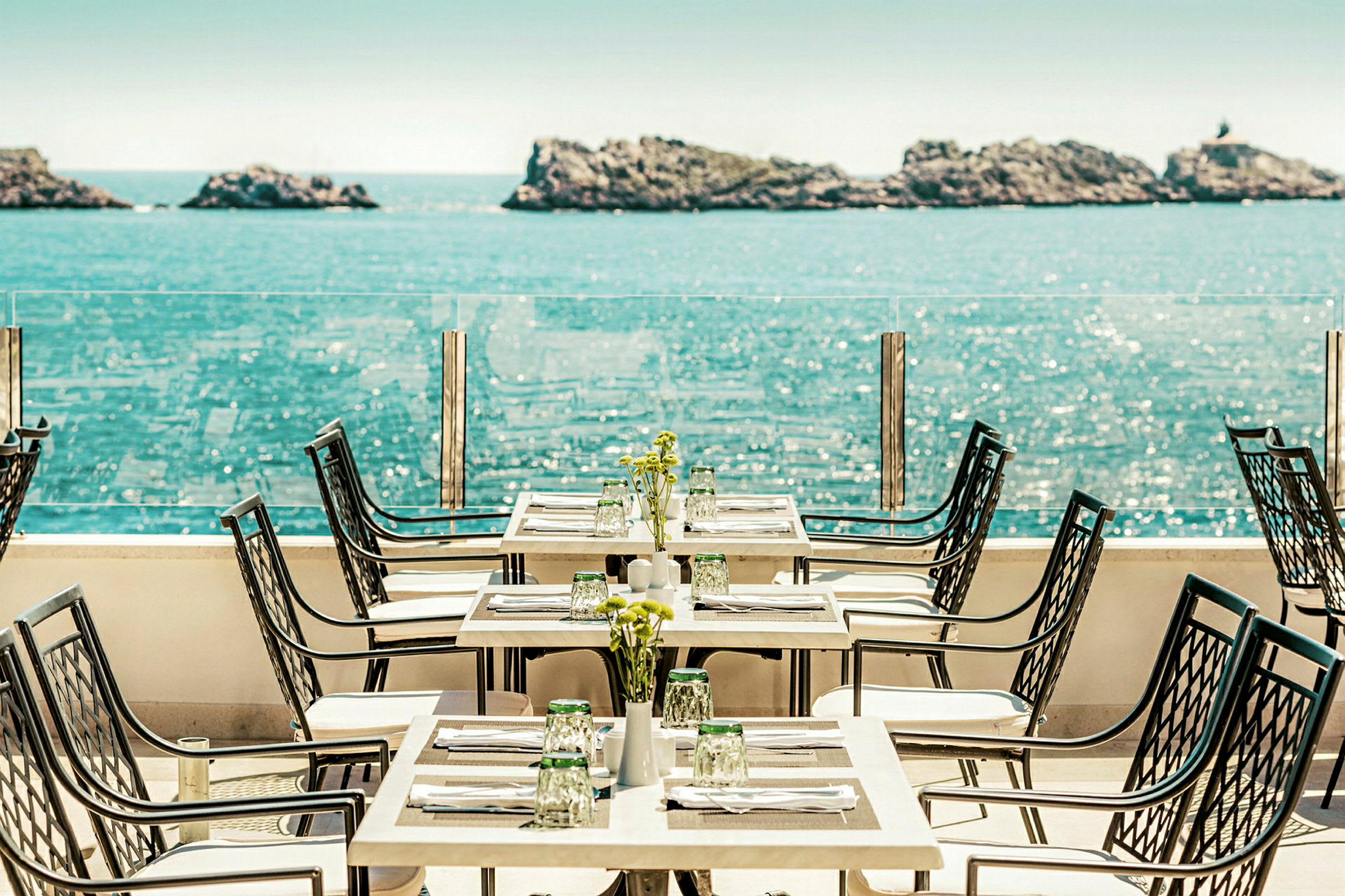 En restaurantterrasse med havudsigt, borde og stole er opstillet med borddækning. (AI-genereret tekst)