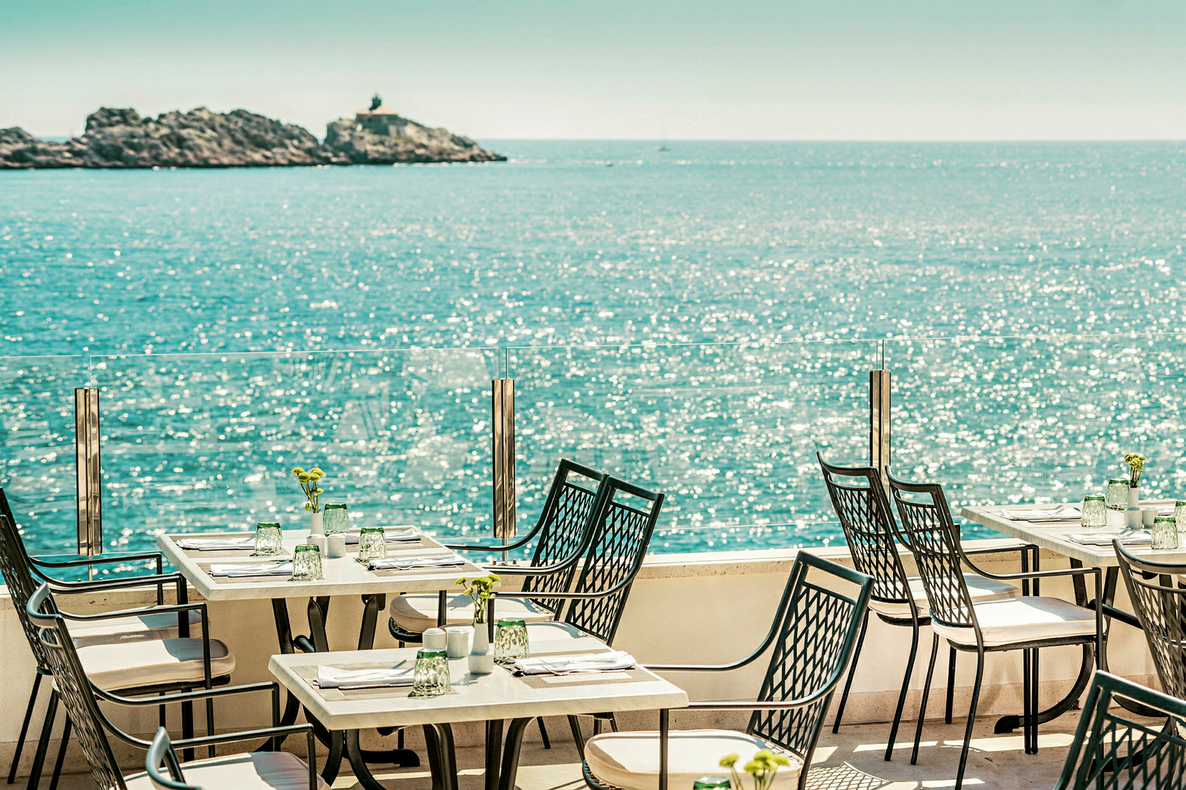 Udendørs serveringsområde med borde og stole ved havet, med udsigt til en klippe i baggrunden. (AI-genereret tekst)
