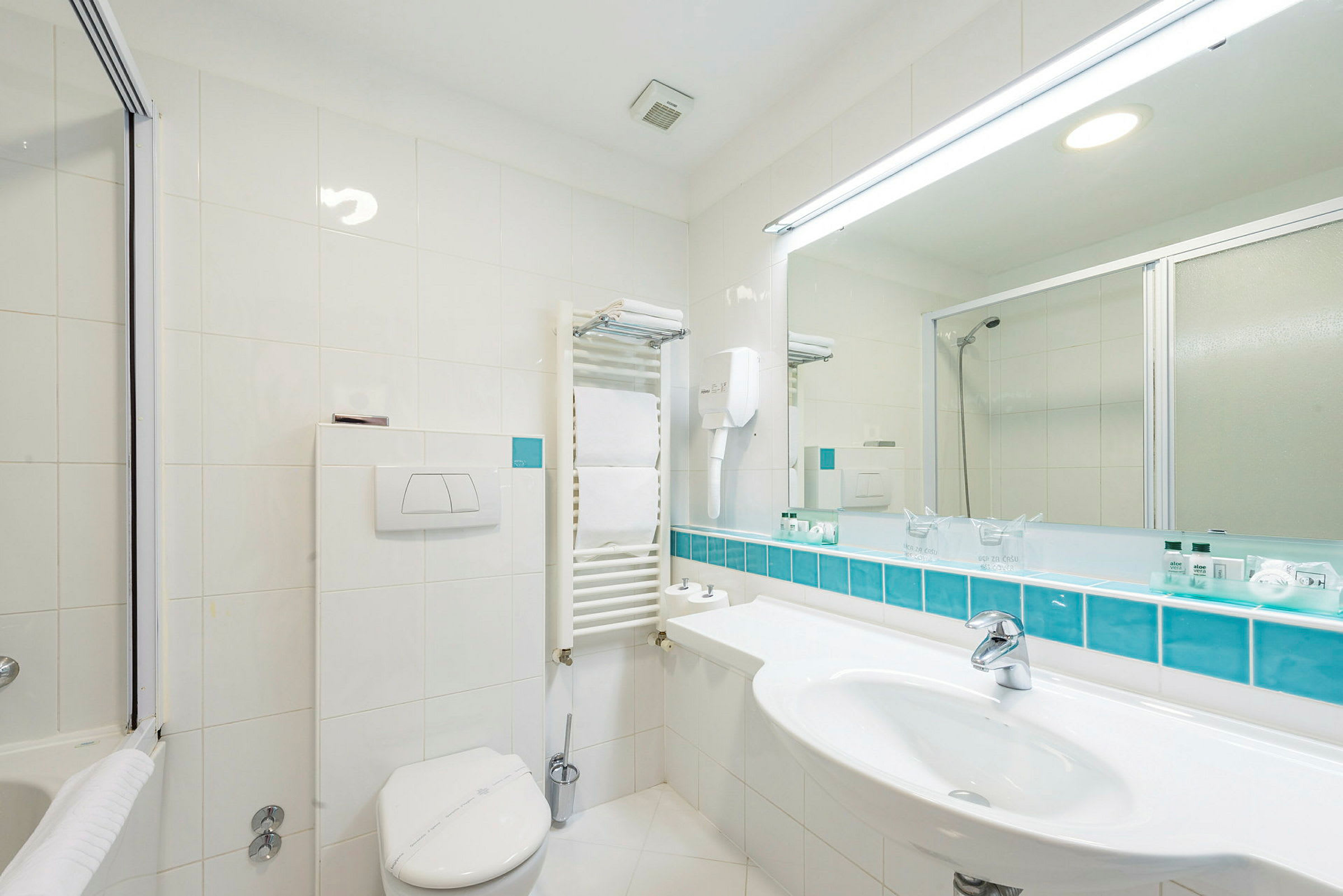 Et moderne badeværelse med hvide flisebeklædte vægge, en håndvask med spejl over, toilet og brusekabine bag en glasdør. (AI-genereret tekst)