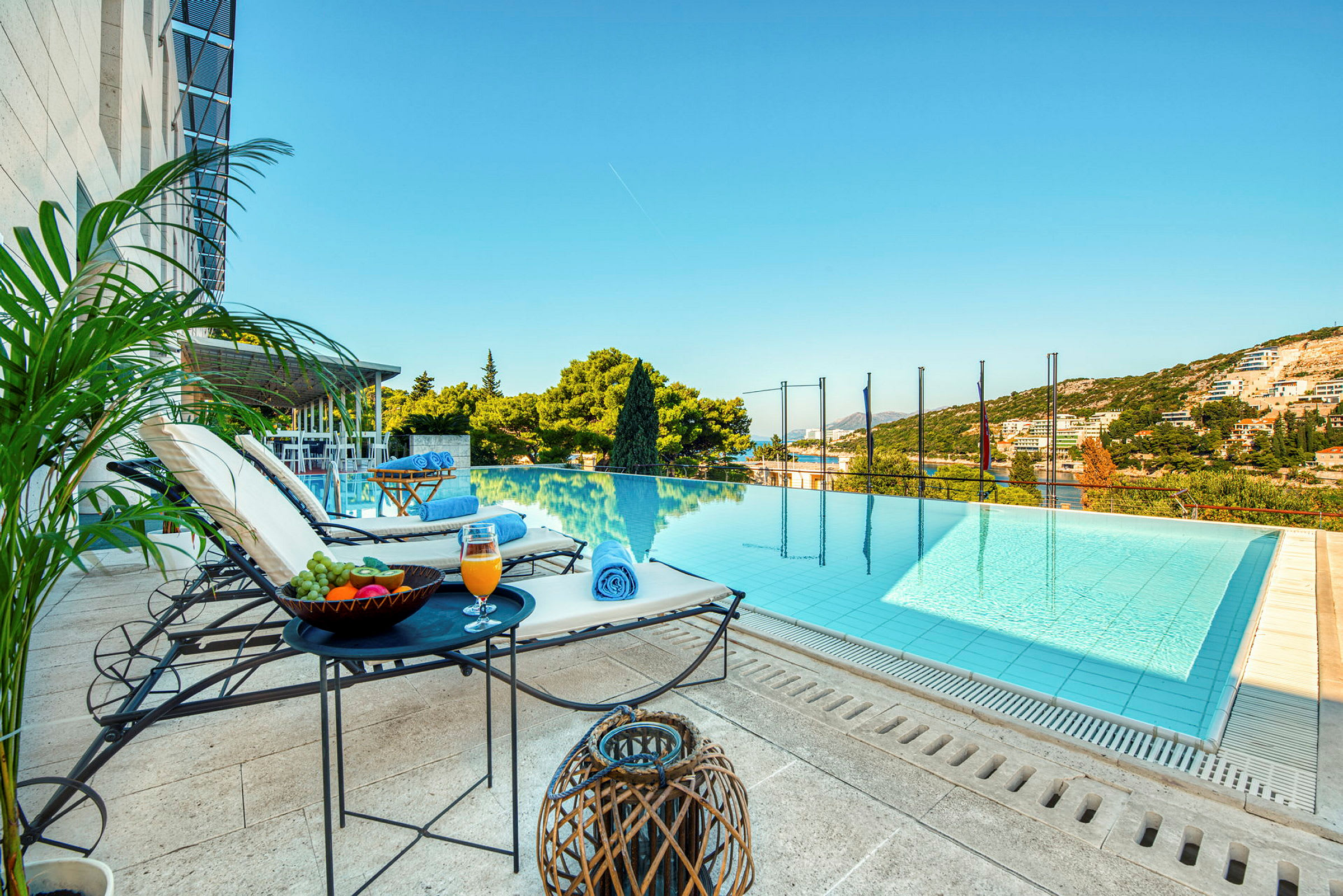 En udsigt over en infinitypool med liggestole, borde med frugt og drikke ved siden af, samt grønne omgivelser og himmel i baggrunden. (AI-genereret tekst)