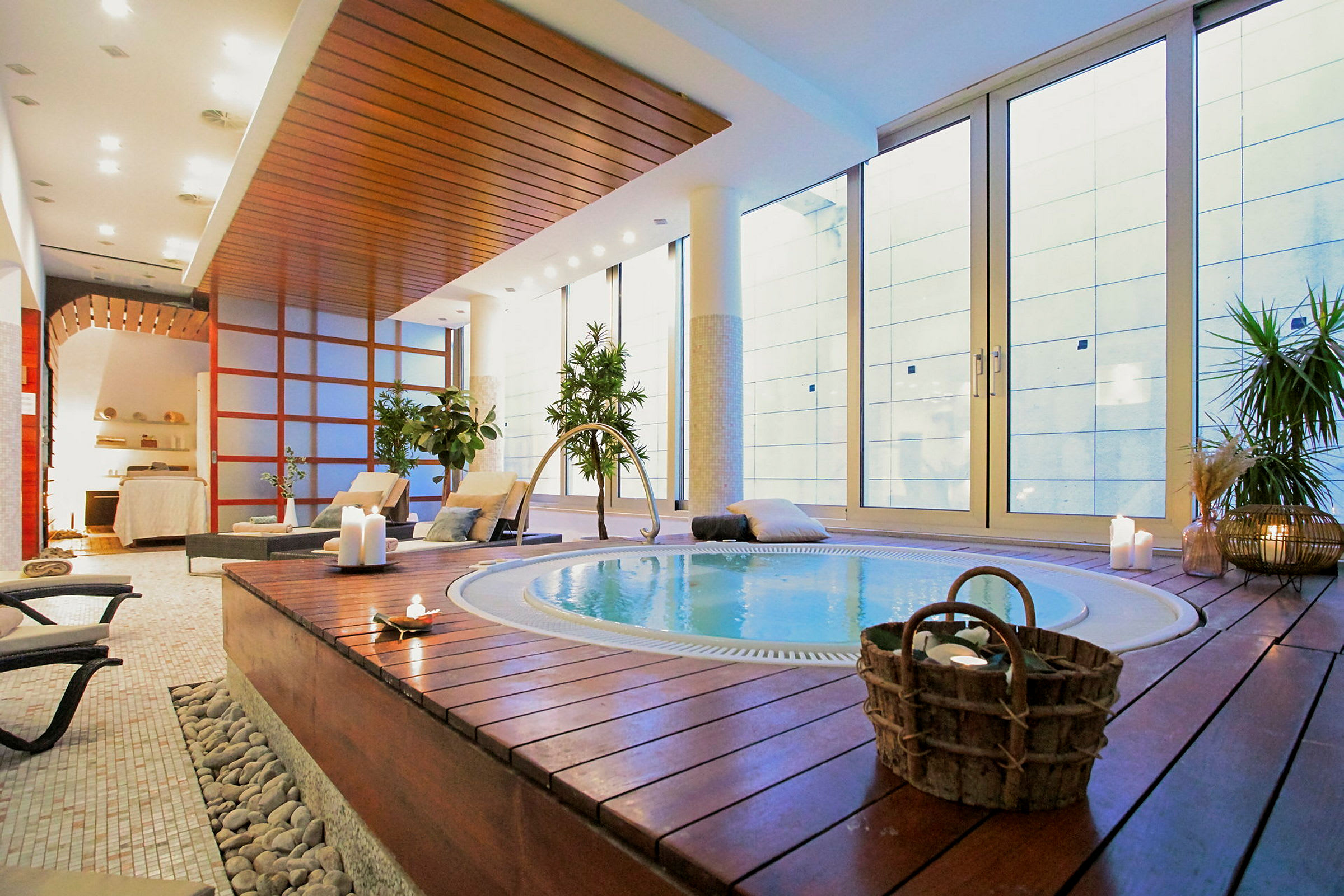 Et elegant spa med en jacuzzi omgivet af træpaneler, stole og levende planter. Store glasvægge giver naturligt lys. (AI-genereret tekst)