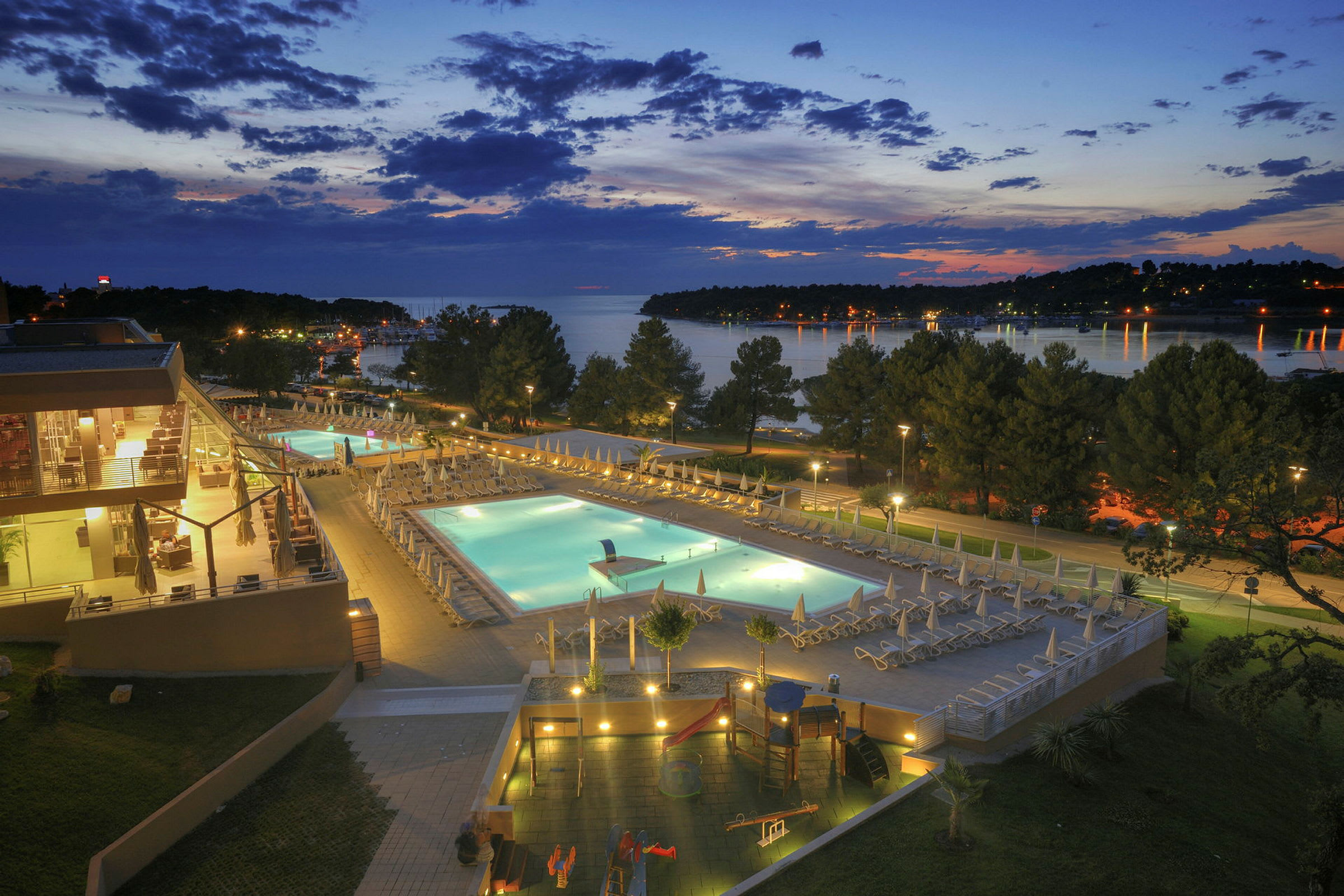 En aftenudsigt over et hotelområde med oplyste swimmingpools, omkringliggende terrasser og træer, med en sø i baggrunden. (AI-genereret tekst)