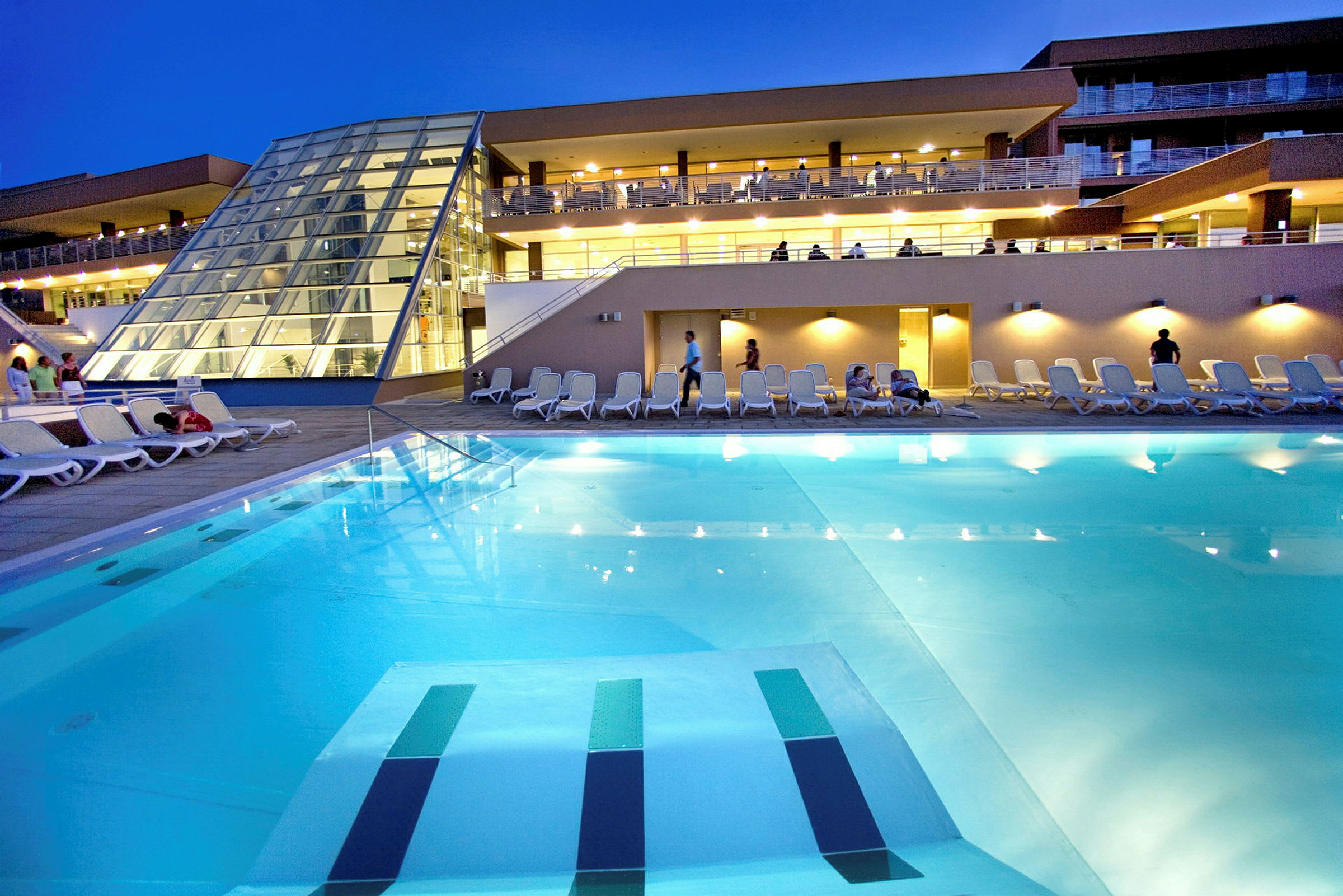 Et aftenbillede af en hotelpool med liggestole omkring og en moderne bygning i baggrunden. (AI-genereret tekst)