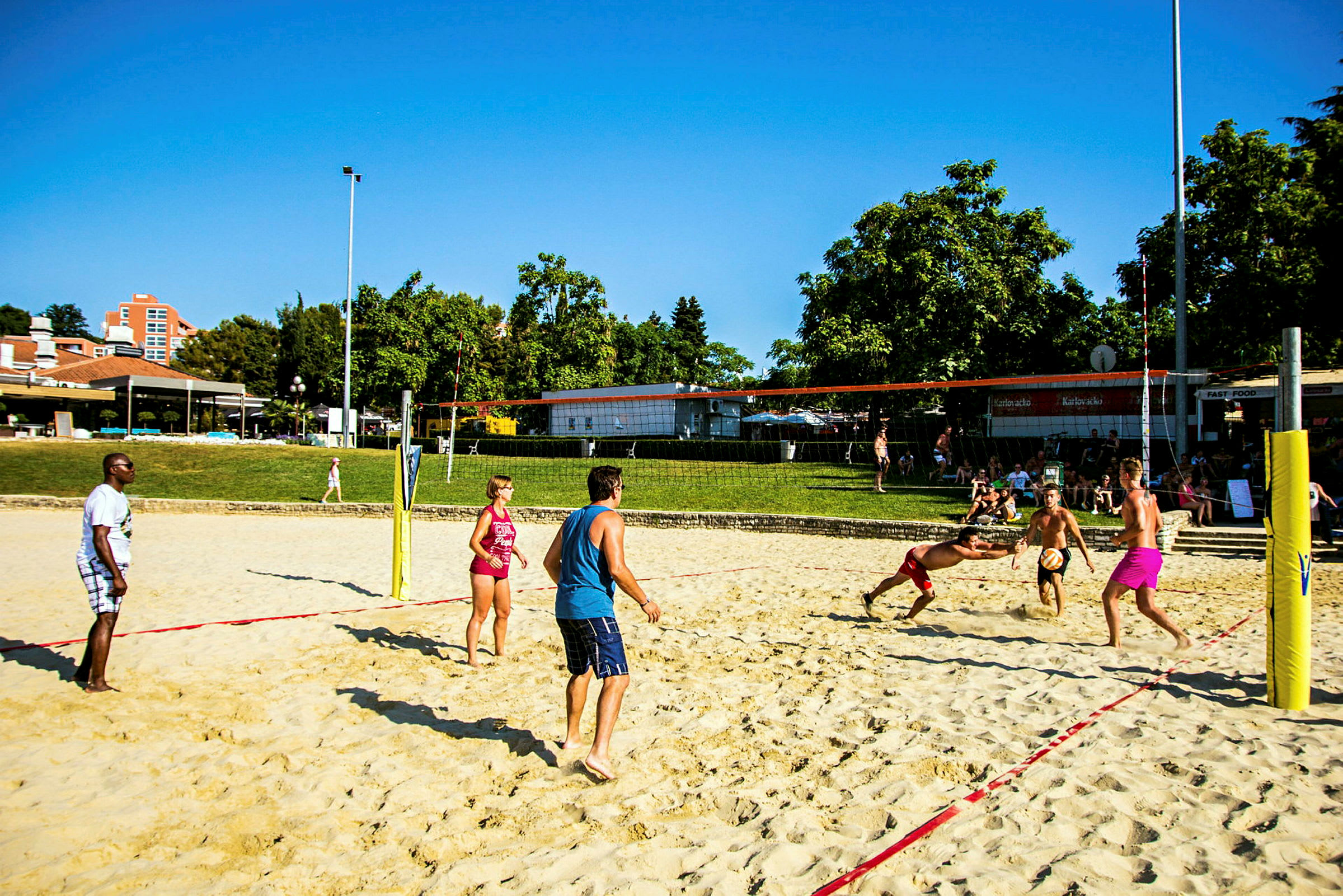 Folk spiller beachvolley på en sandbane i et udendørs område med træer og bygninger i baggrunden. (AI-genereret tekst)