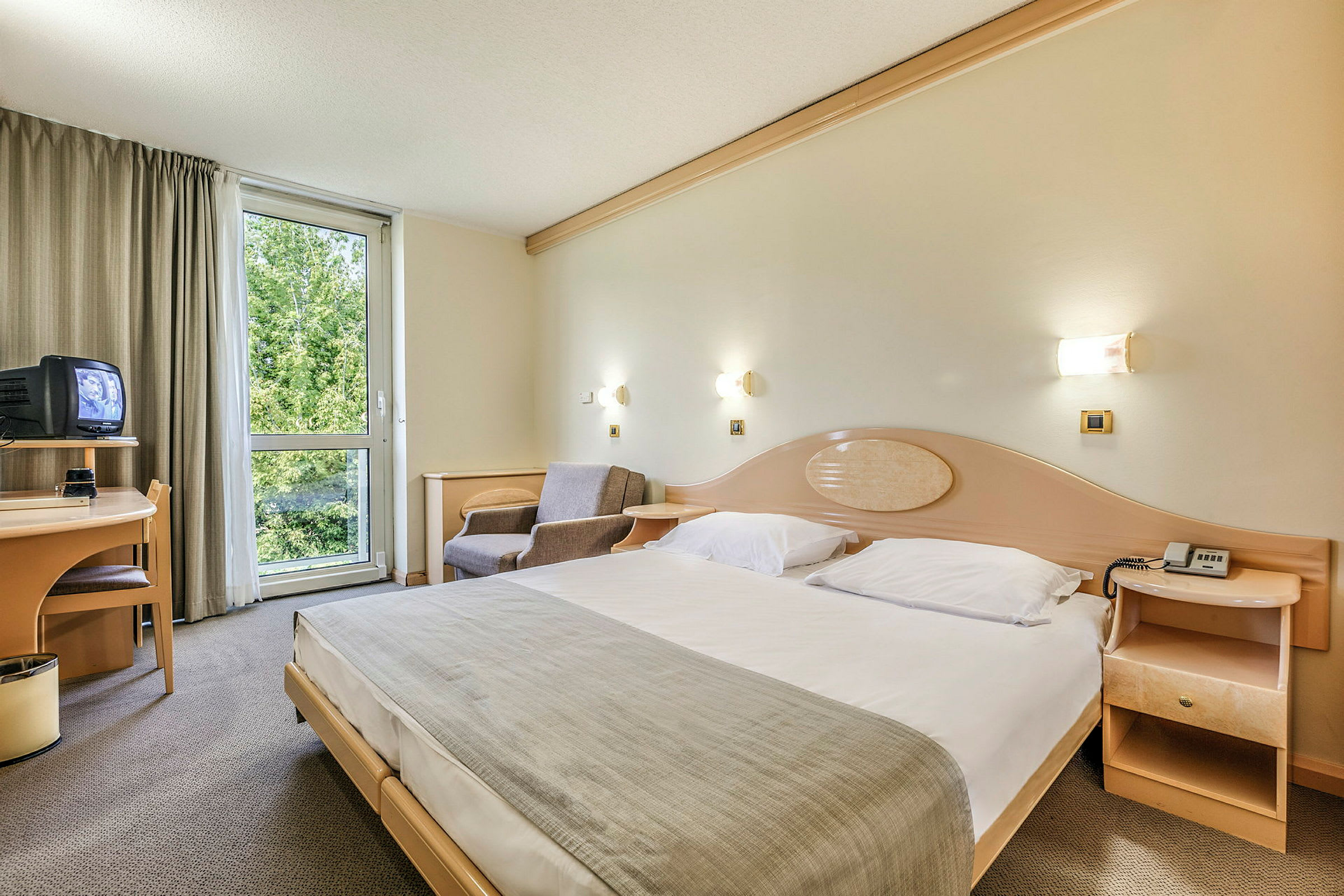 Hotelværelse med en stor seng, en lænestol, et skrivebord med et TV og et vindue med udsigt til grønne områder. (AI-genereret tekst)