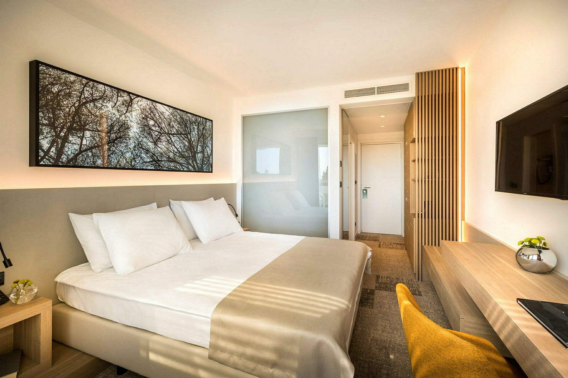 Et moderne hotelværelse med en stor seng med hvidt sengetøj. Værelset har en minimalistisk indretning med et vægmonteret TV, et skrivebord og en stol. (AI-genereret tekst)